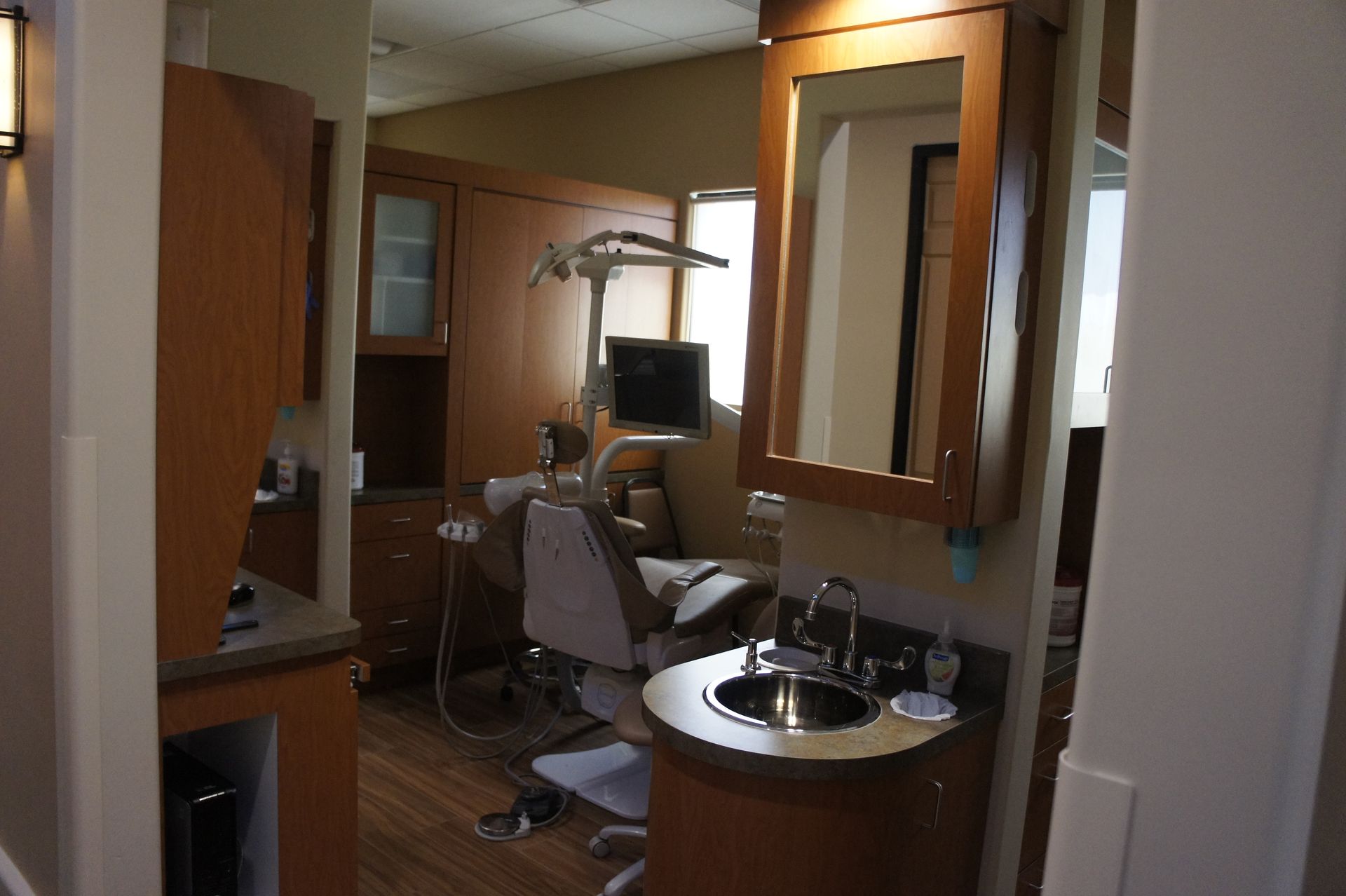 A dentist's office with a chair, cabinets, sink, and window. Wooden cabinets and light colors.