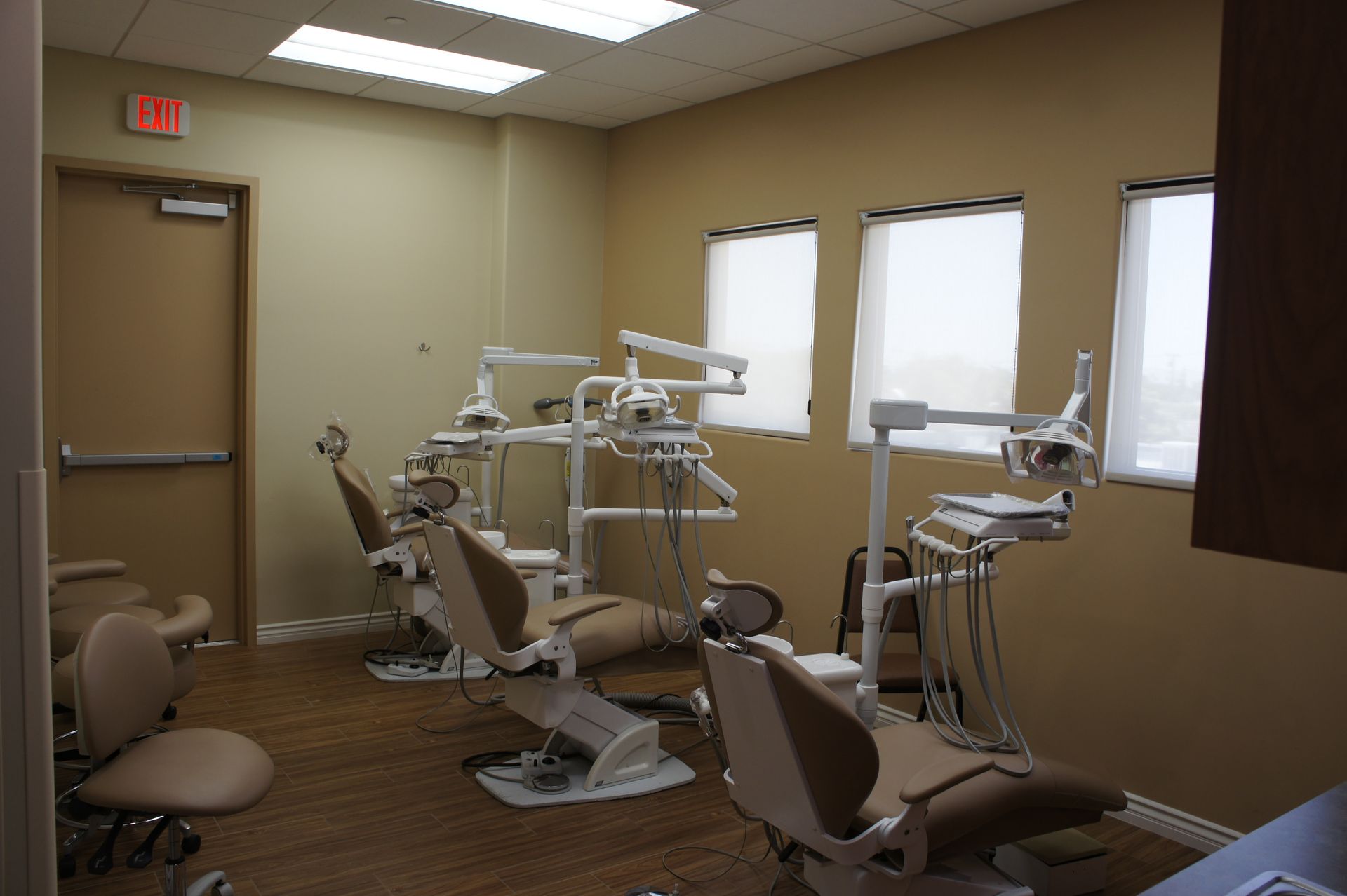 Dentist office with several dental chairs, equipment, and windows; beige walls and brown flooring.