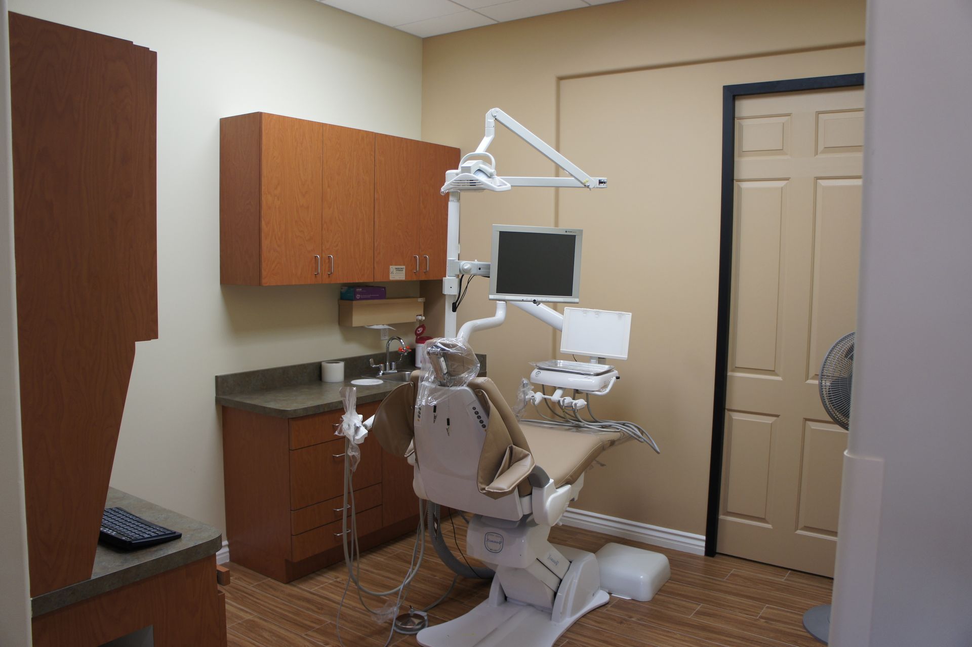 Dental office room with chair, equipment, cabinets, and door. Beige and brown tones.