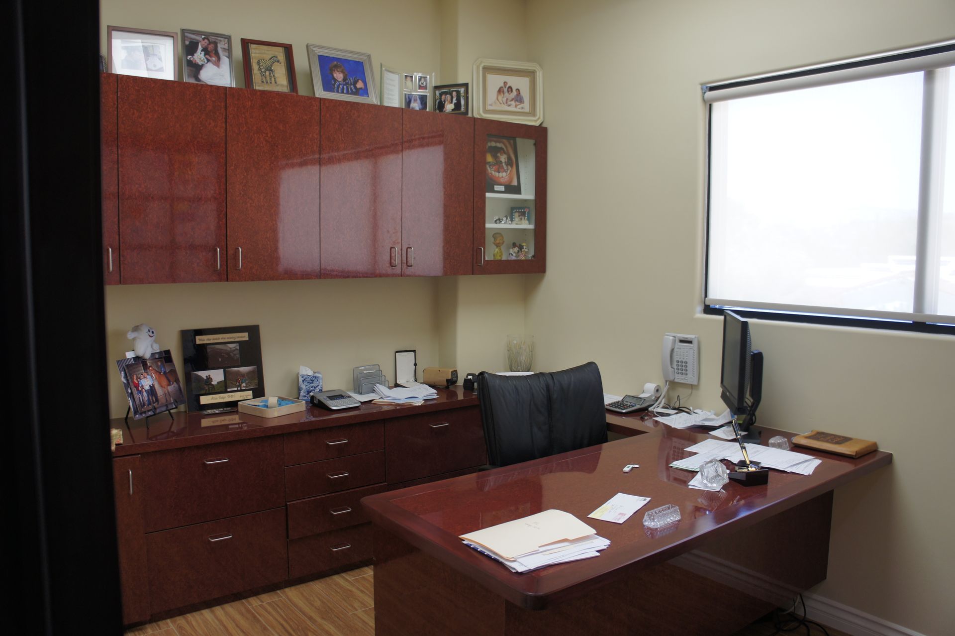 Office with dark wooden desk, cabinets, and chair. Window with white shade. Cluttered desk surface.