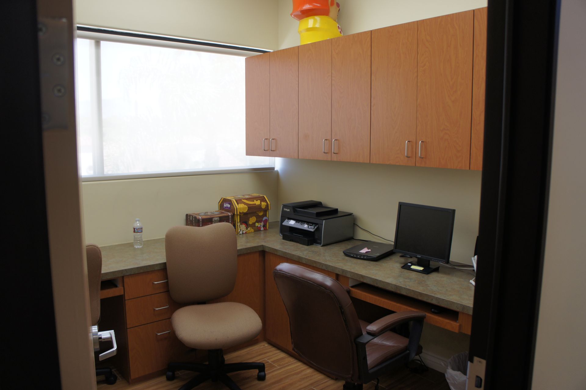Small office with two desks. One with a computer and printer, cabinets overhead.
