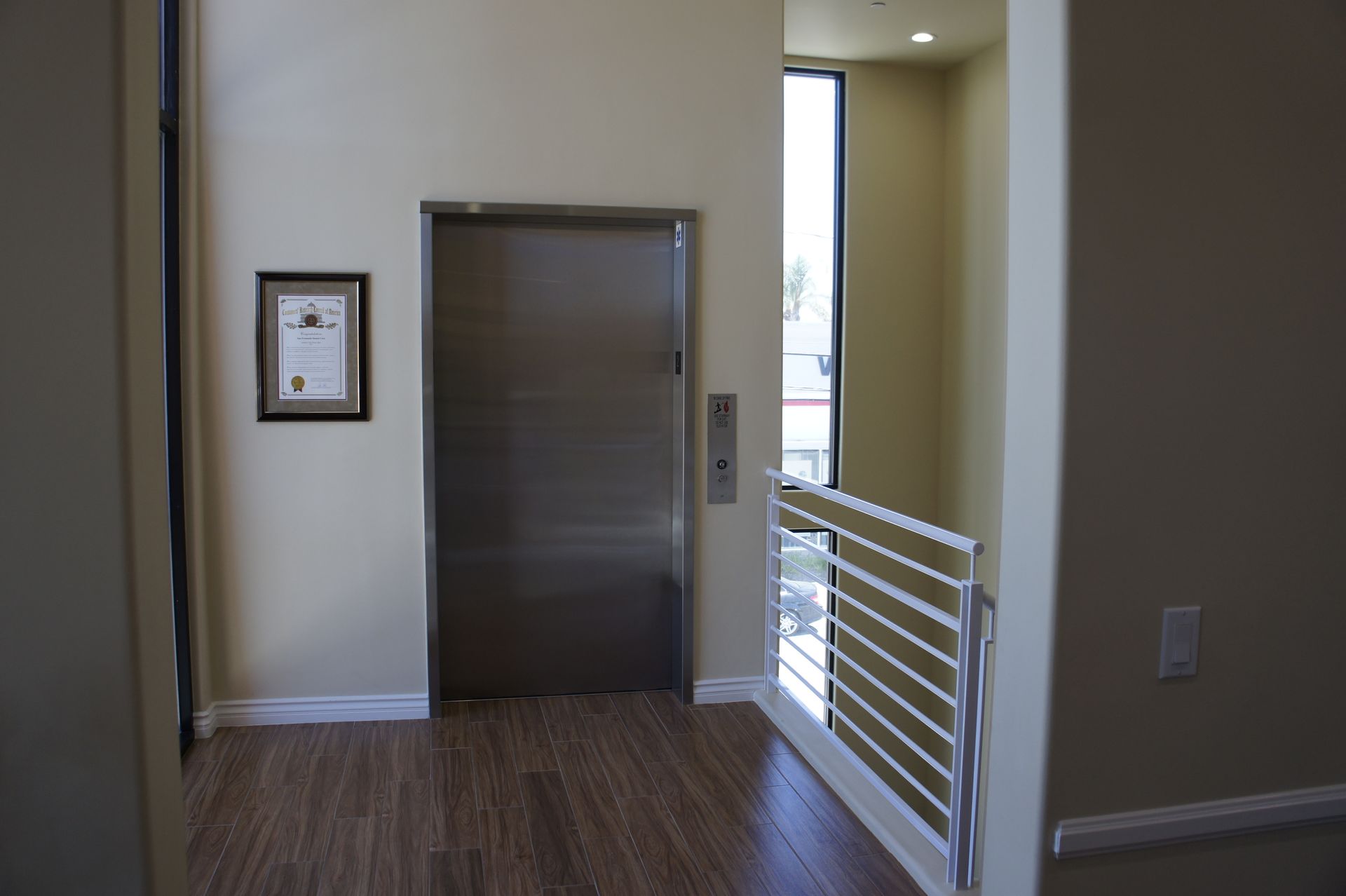 Elevator with closed metal door, button panel, framed document on wall, and a railing to the right.
