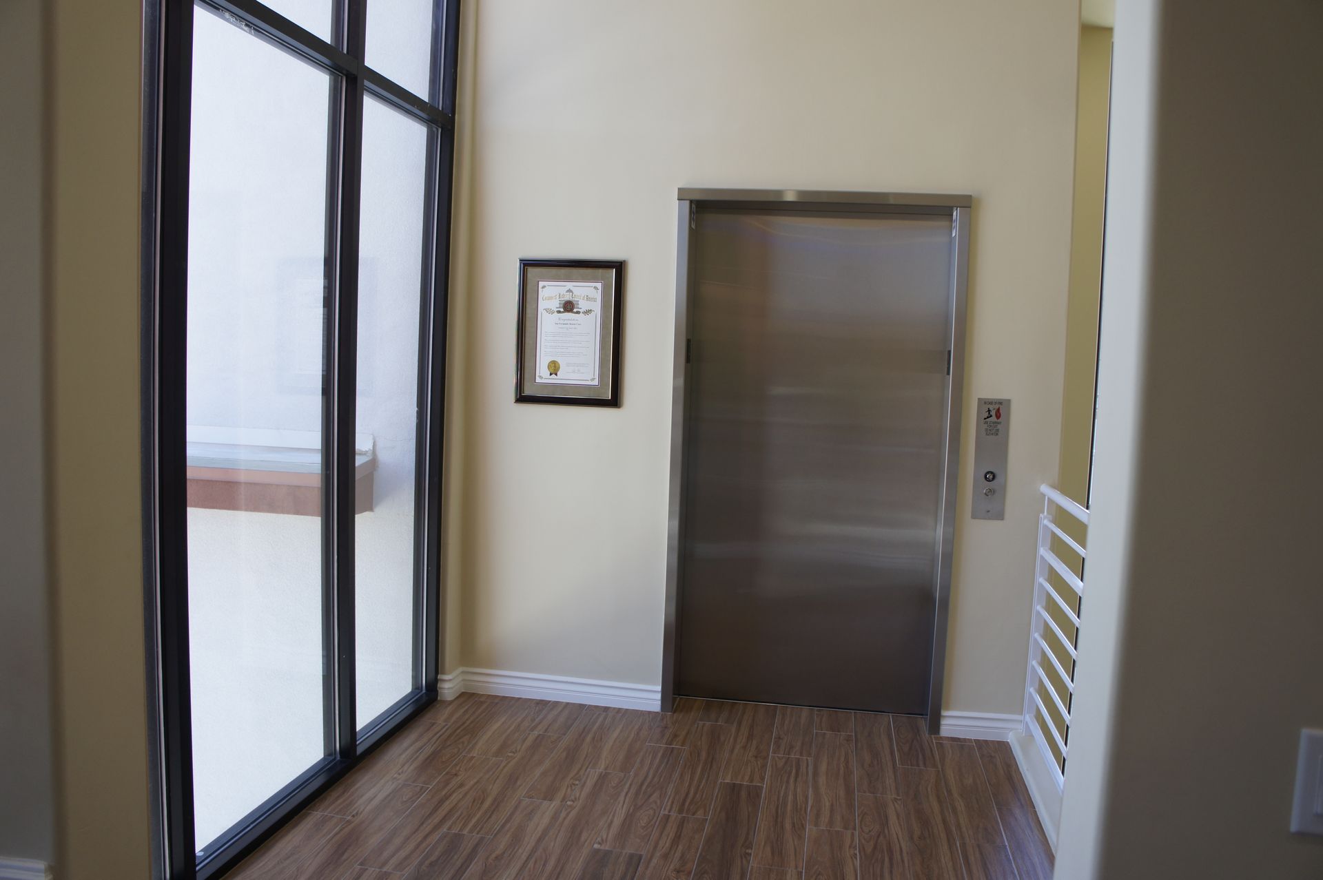 Interior view of a building with a stainless steel elevator door, wooden floor, and large window.