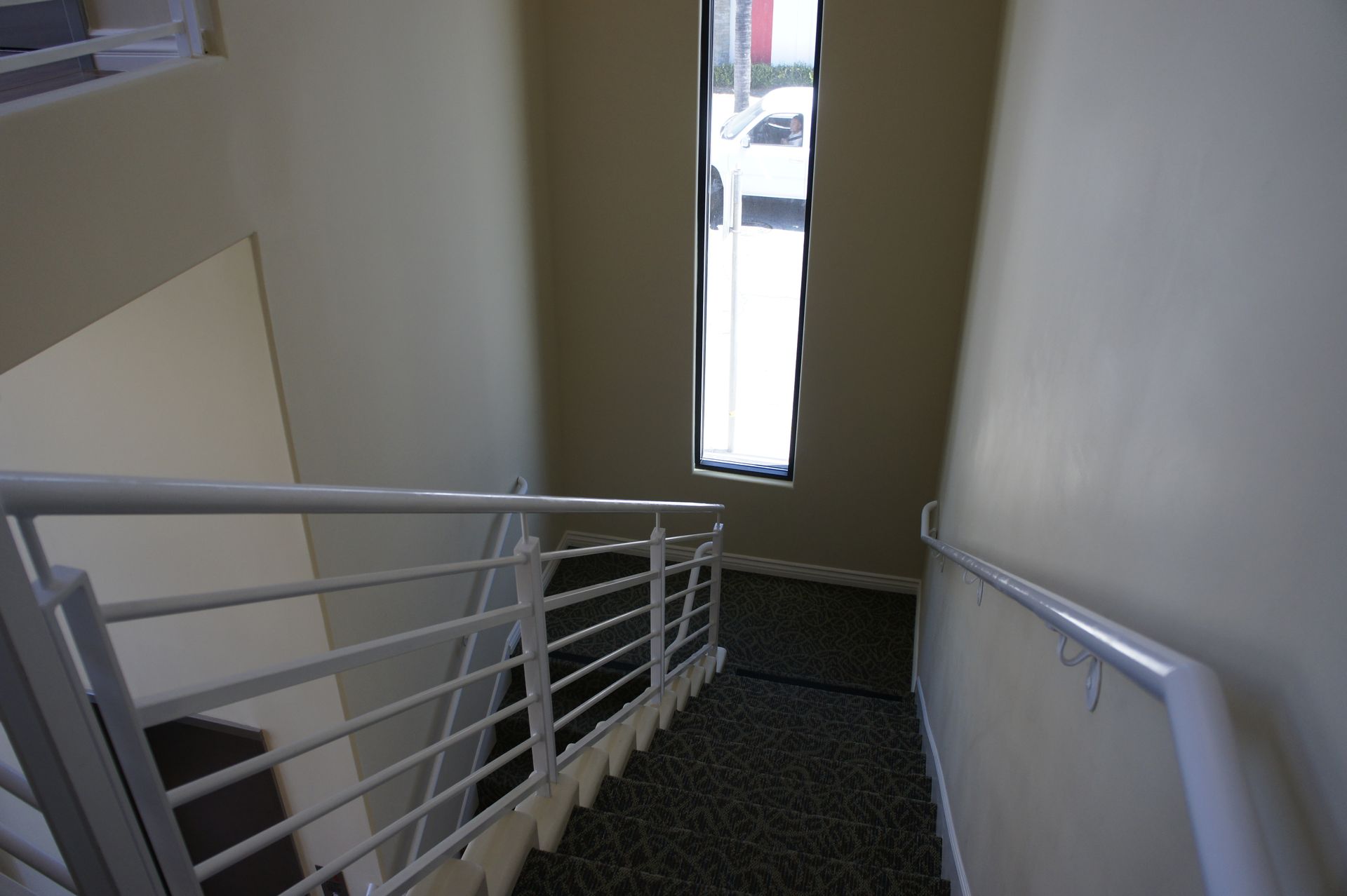 Stairwell with white railings, beige walls, and a tall window at the bottom, natural light.