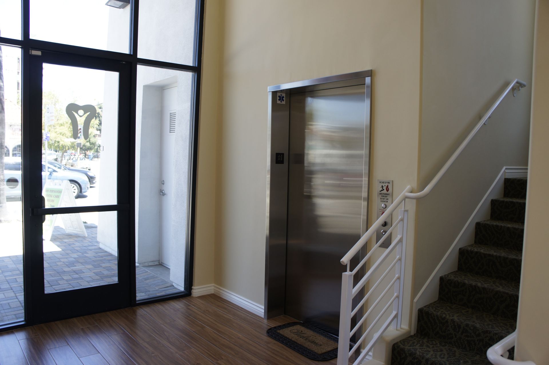 Entrance lobby with elevator, stairs, and glass door. Bright interior, neutral walls, and black door frame.