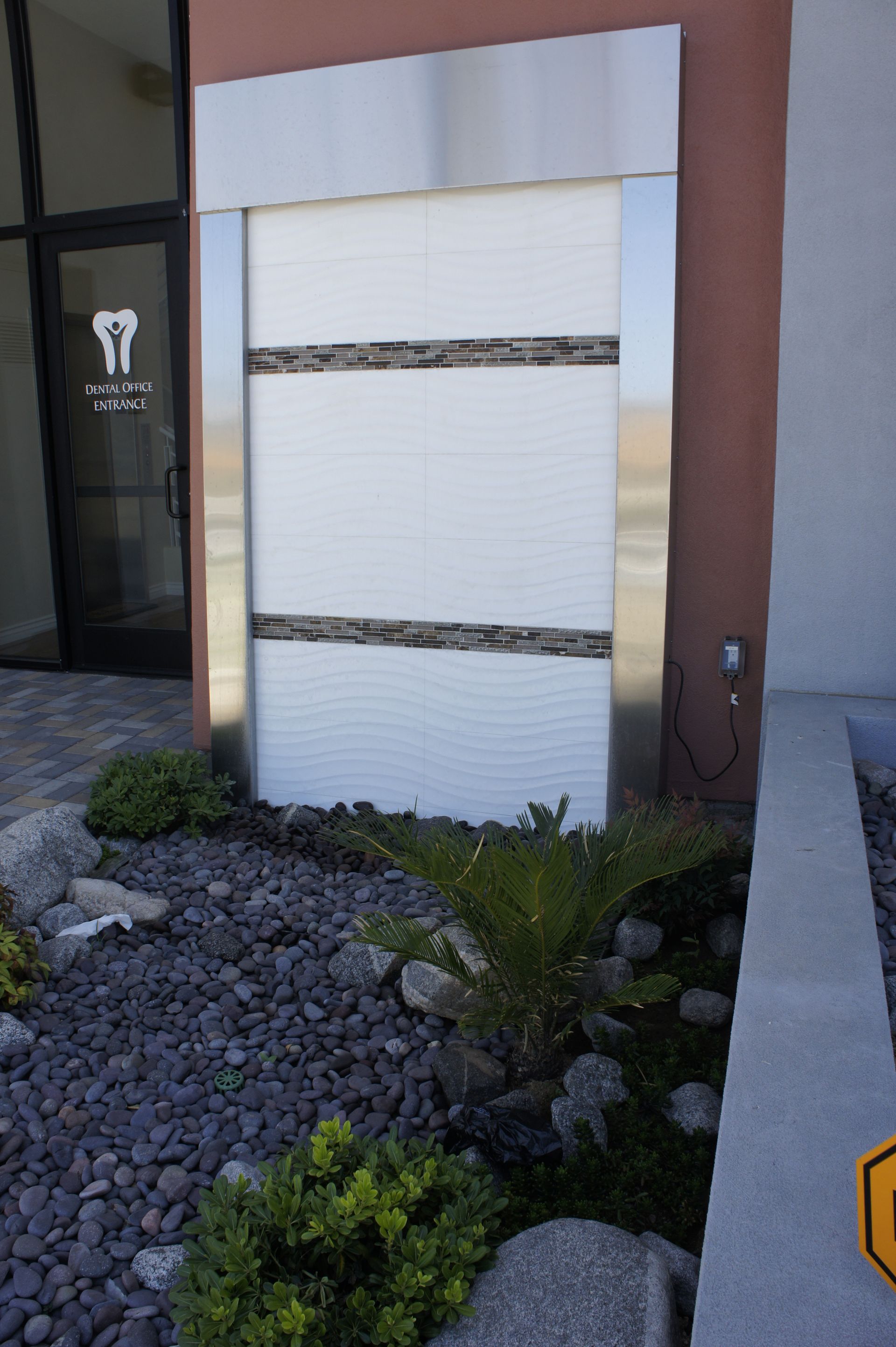 Water fountain outside a dental office, white textured tile with dark stone accents, surrounded by landscaping.
