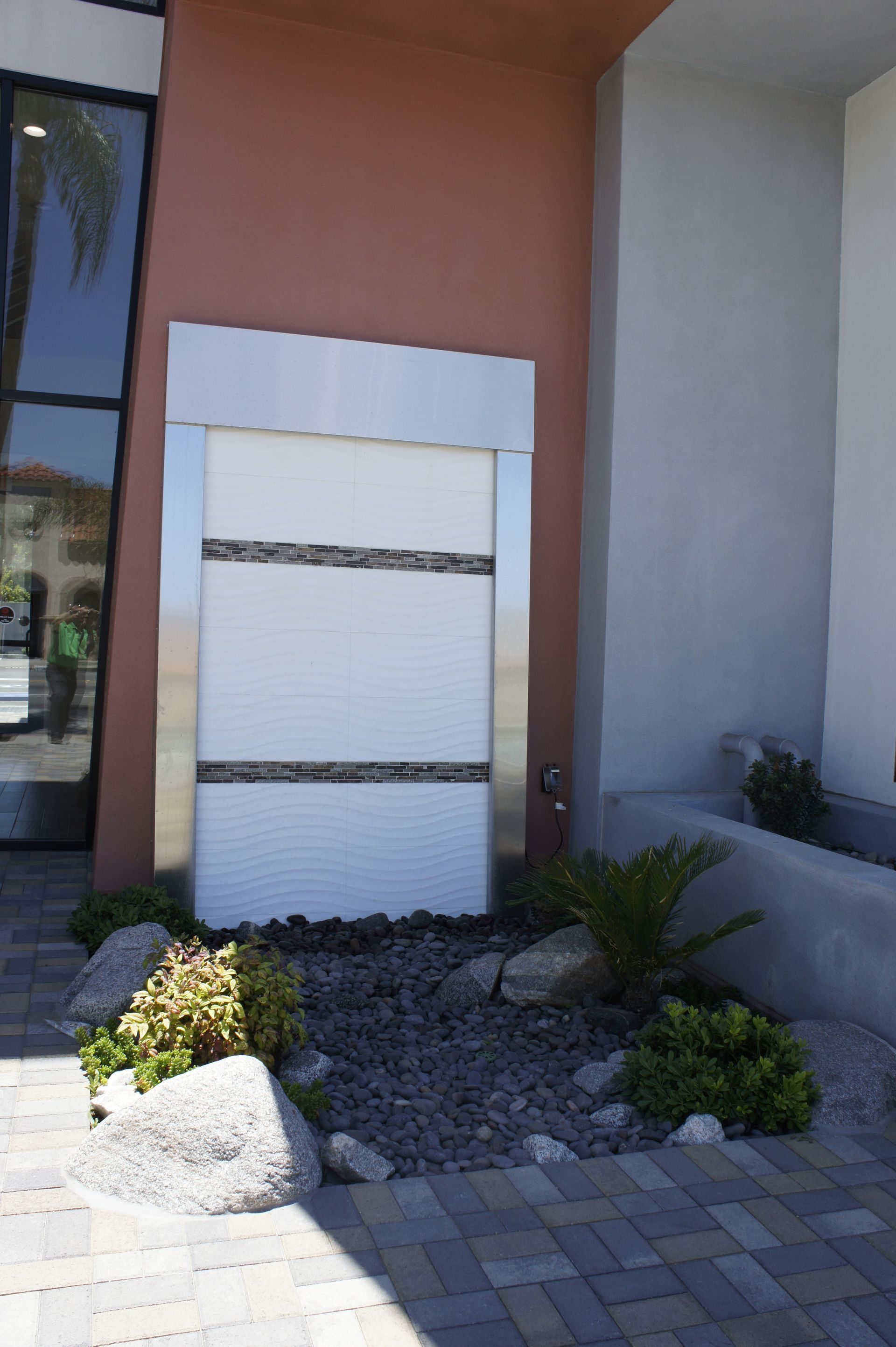 Modern outdoor water fountain with white tiled face, gray stone border, and landscaping.