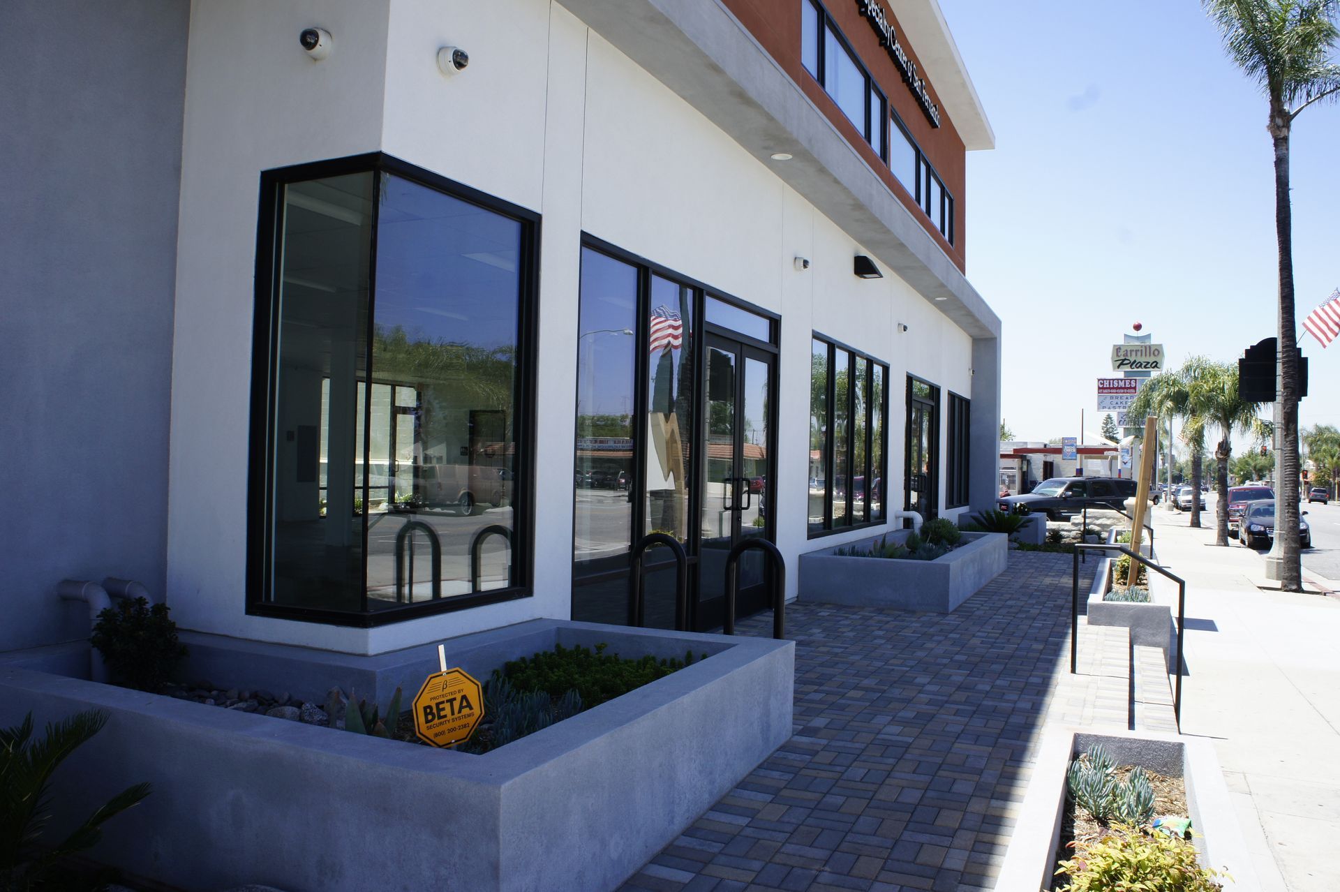 Exterior of a modern building with large windows, concrete planters, and a brick walkway, on a sunny day.