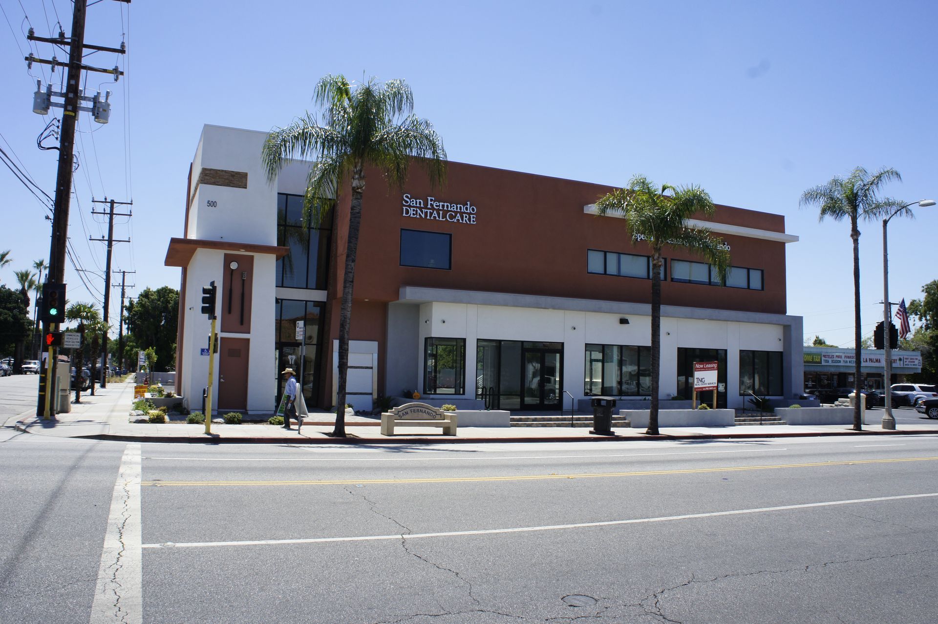Modern, two-story building with white and brown facade on a sunny street corner. Palm trees in front.