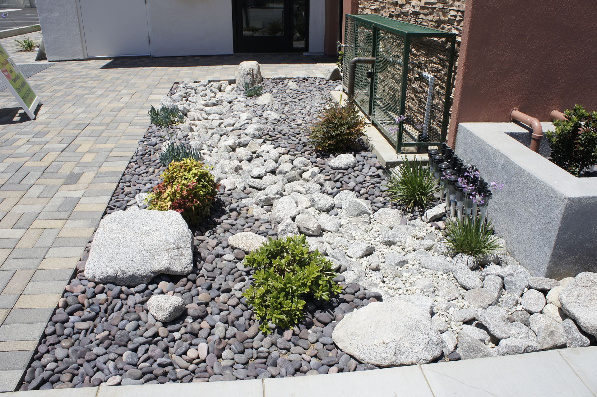 Rock garden with various plants and stones in front of a building with brick pavers.