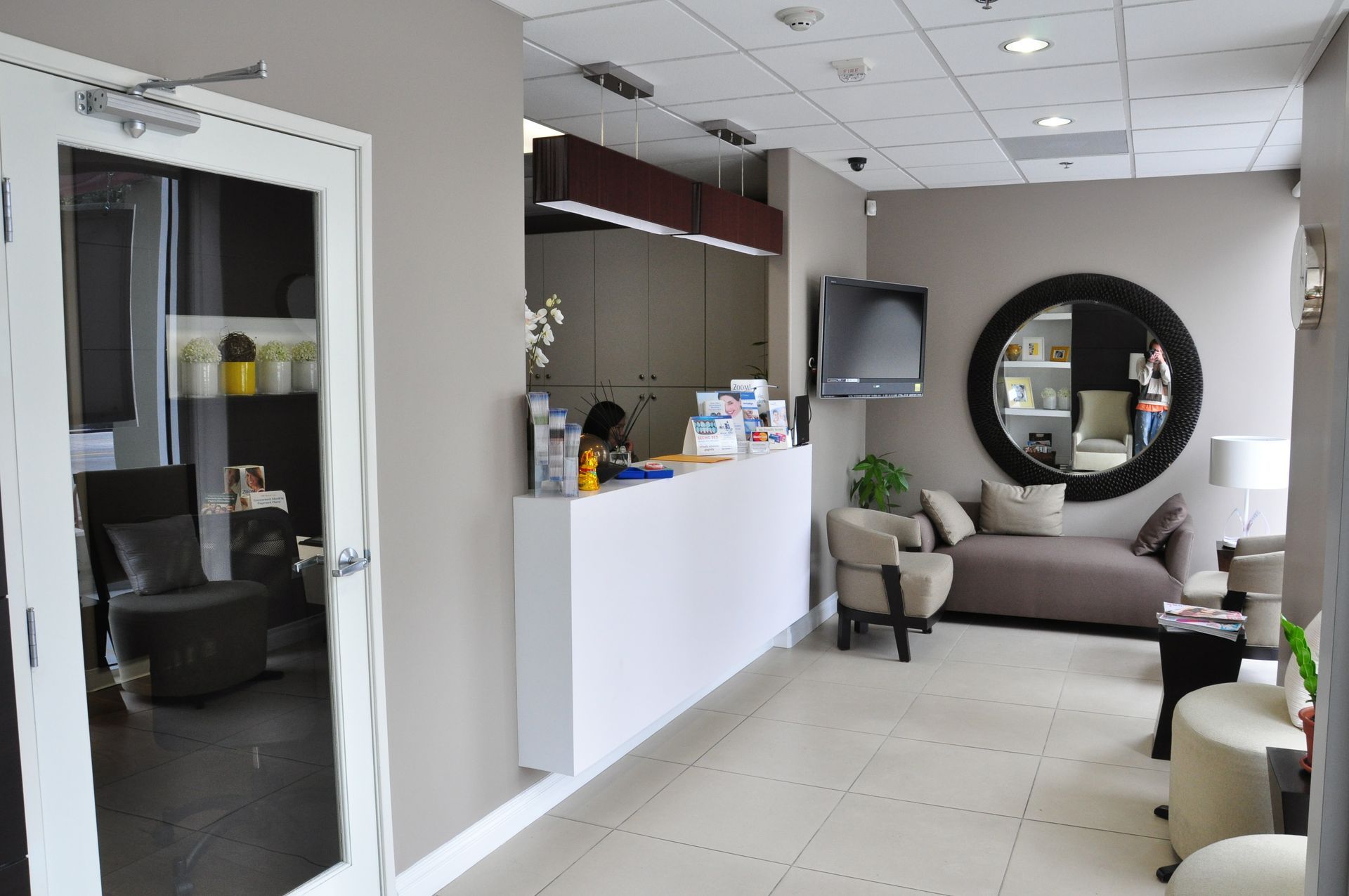 Reception area with white desk, beige walls, seating, and large mirror.