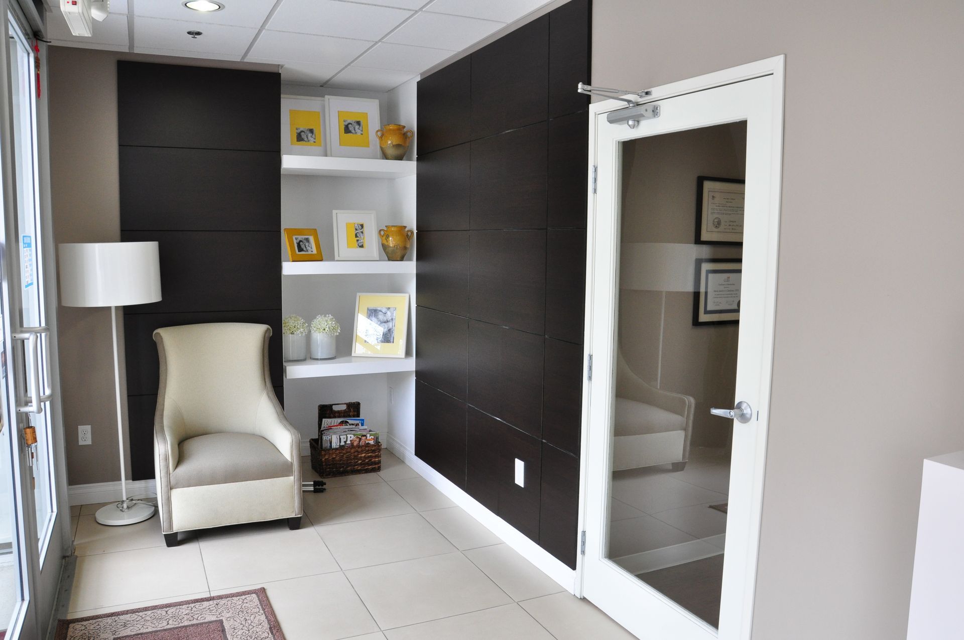 Waiting room with chair, framed art, and shelves against a dark wall, with a glass door to another room.