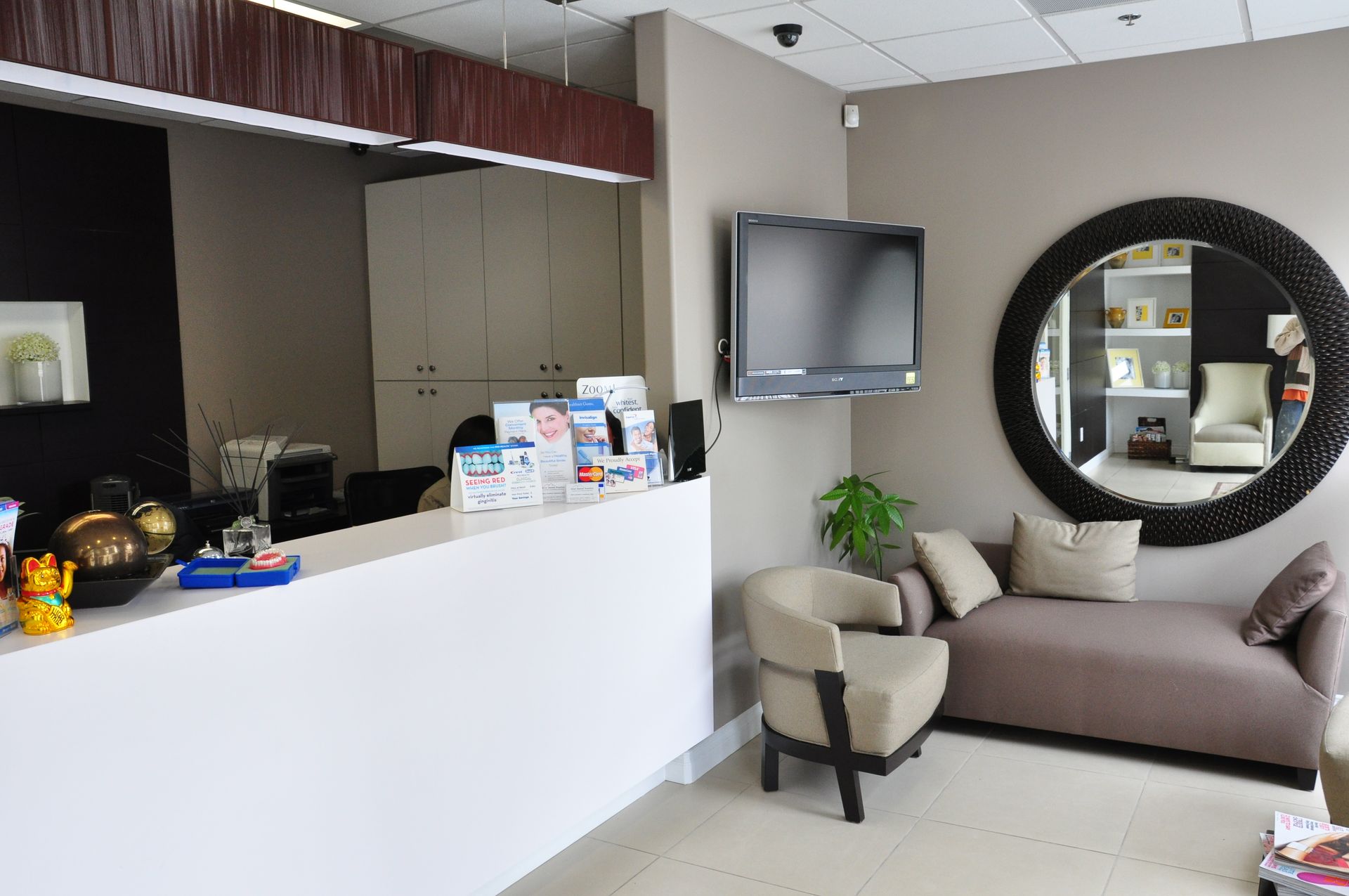 Reception area with a white desk, seating, a TV, and a large mirror on a beige wall.