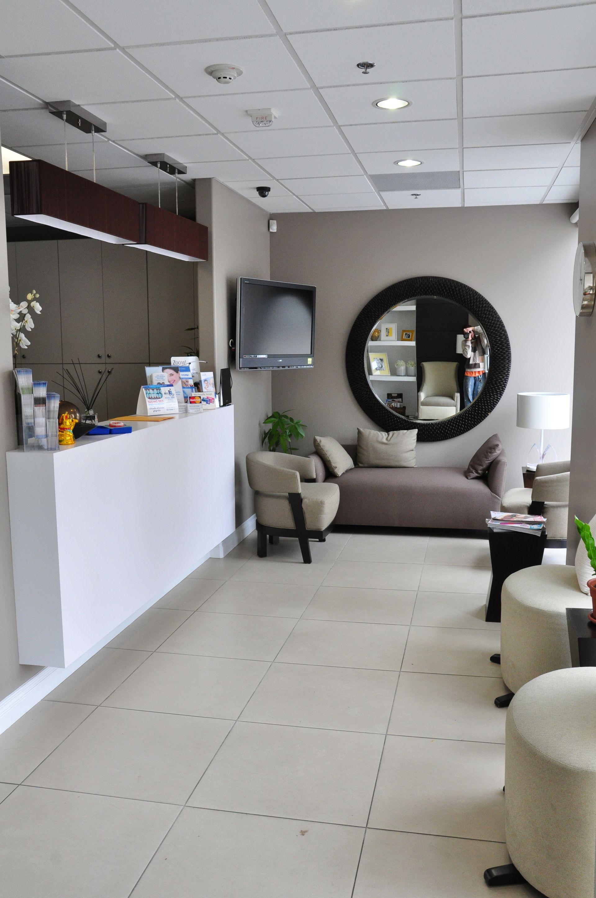 Reception area with white desk, gray walls, sofa, chairs, and a large round mirror.