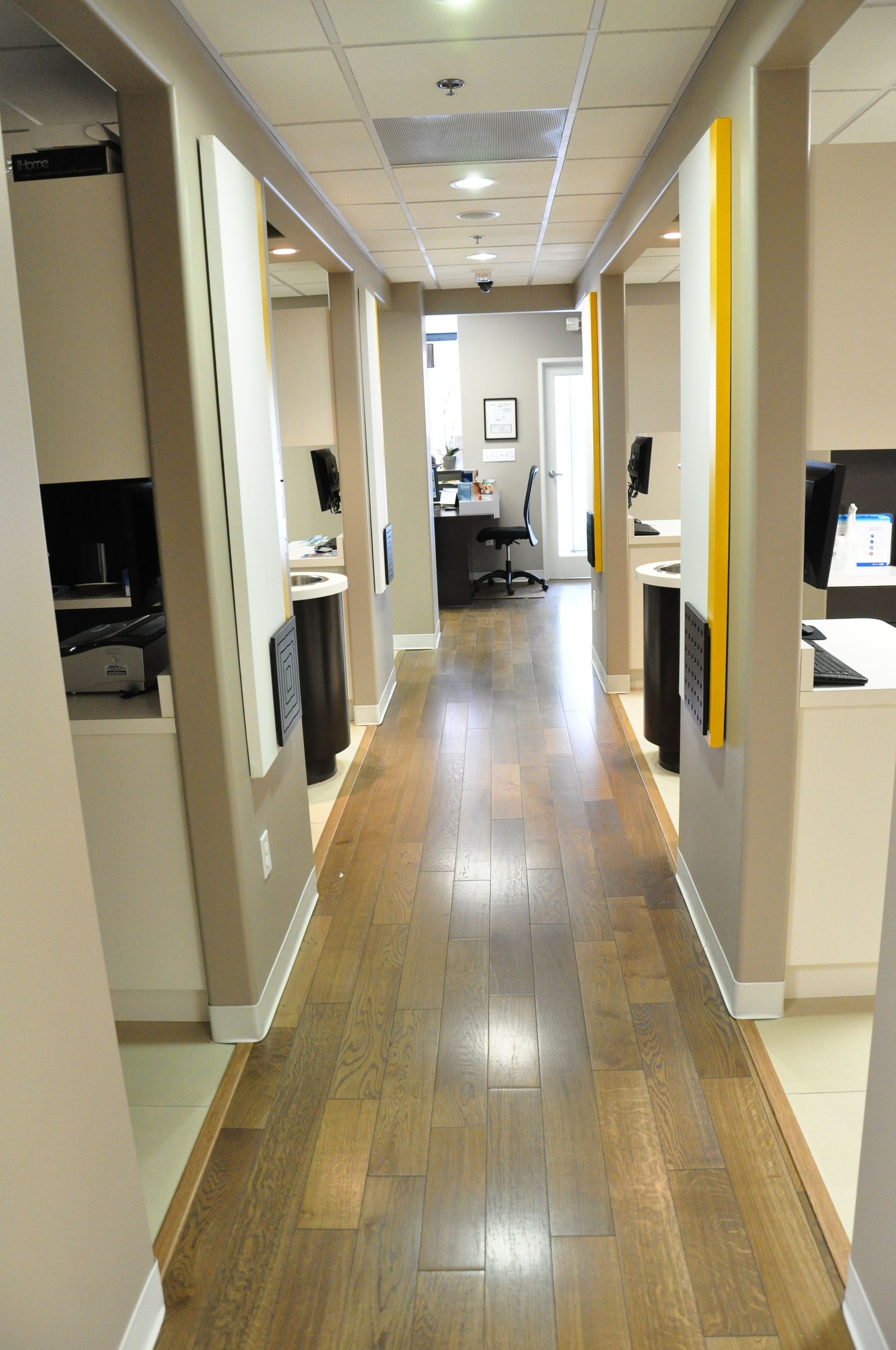 Hallway with dental stations. Wooden floor, tan walls, yellow accents, and computer stations line the sides.