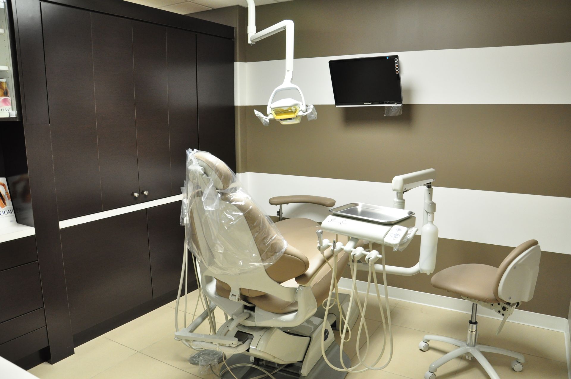 Dentist's office interior with dental chair, brown cabinets, striped wall, and a mounted TV.