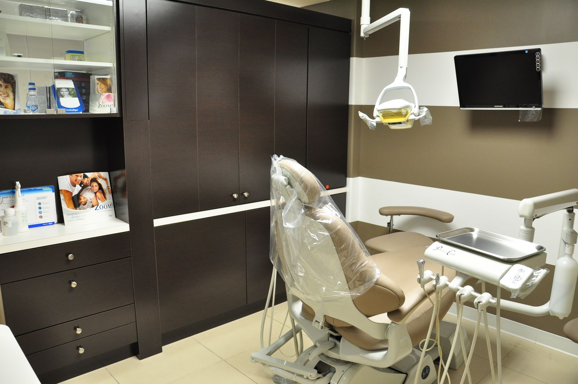 Dental office exam room with chair, equipment, cabinets, and a wall-mounted TV.