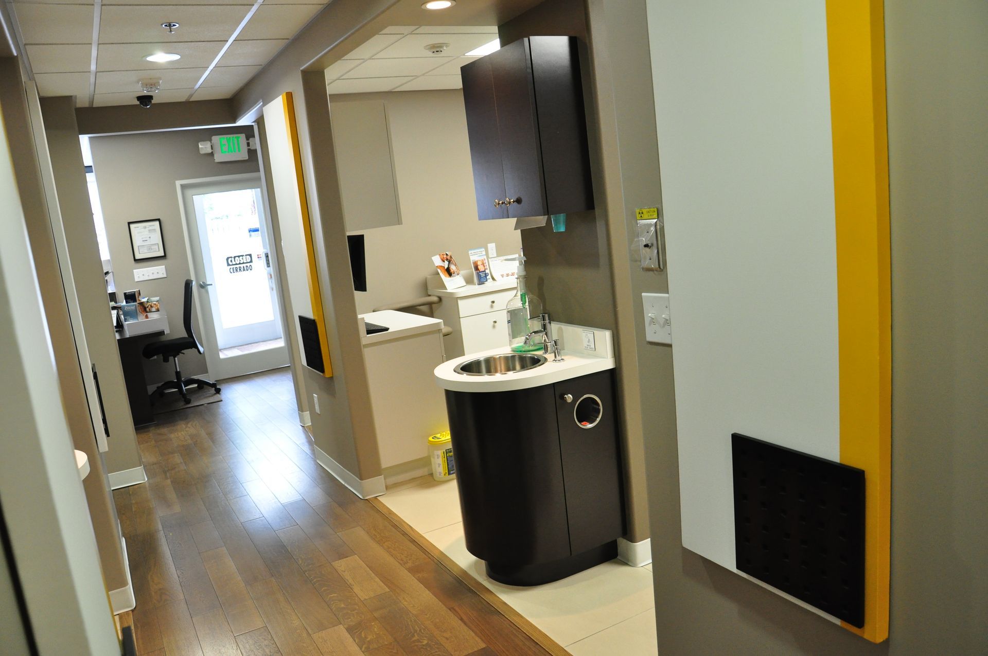 Hallway in a dental office with a sink, cabinets, and a door at the end. Wooden floors and gray walls.