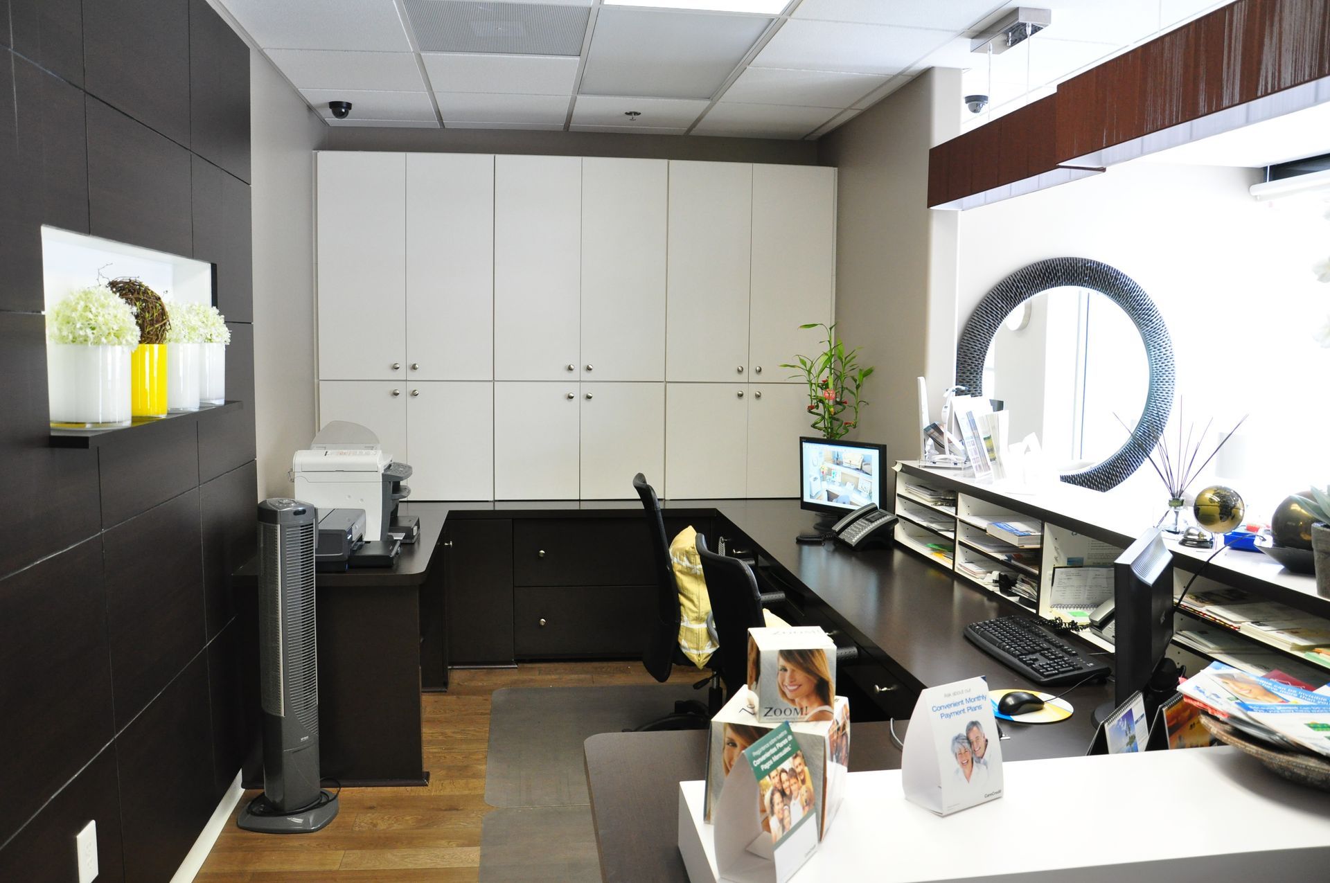 Reception area with dark wood accents, white cabinets, and desk; computer and fan visible.