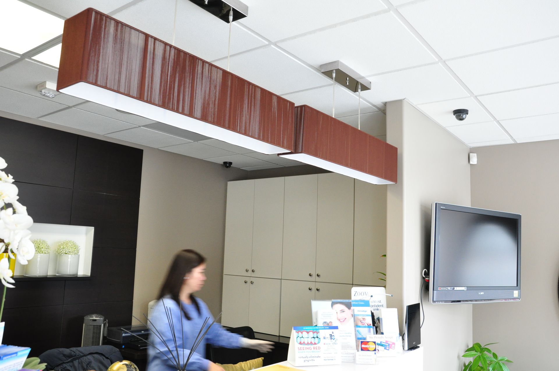 A woman at a reception desk in a medical office with long rectangular pendant lights above.