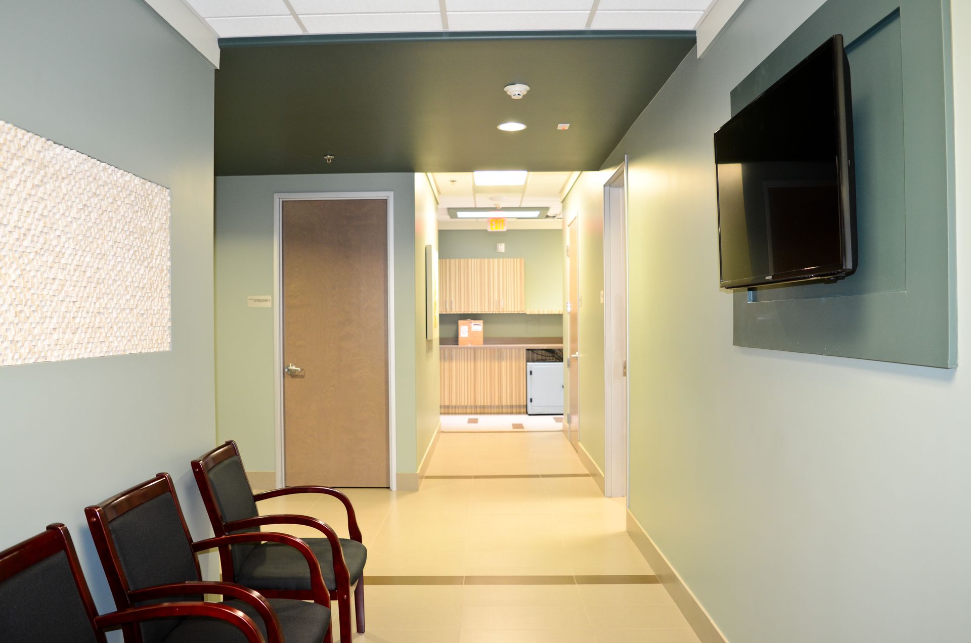 A hallway in a clinic with a row of chairs, a TV, and a door leading to another room.