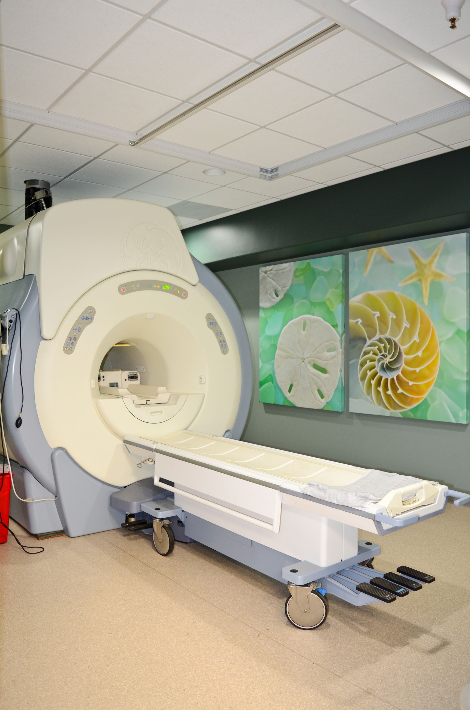 MRI machine in a medical setting. The machine is light blue and white, with a white patient table. Two paintings hang on the wall.