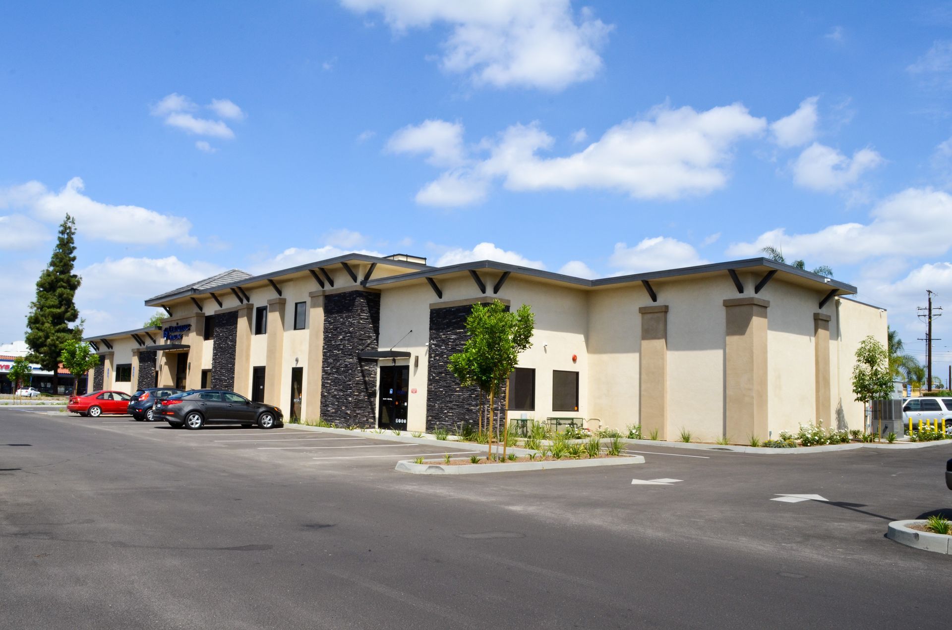 Two-story beige commercial building with a black stone facade and cars parked in front on a sunny day.