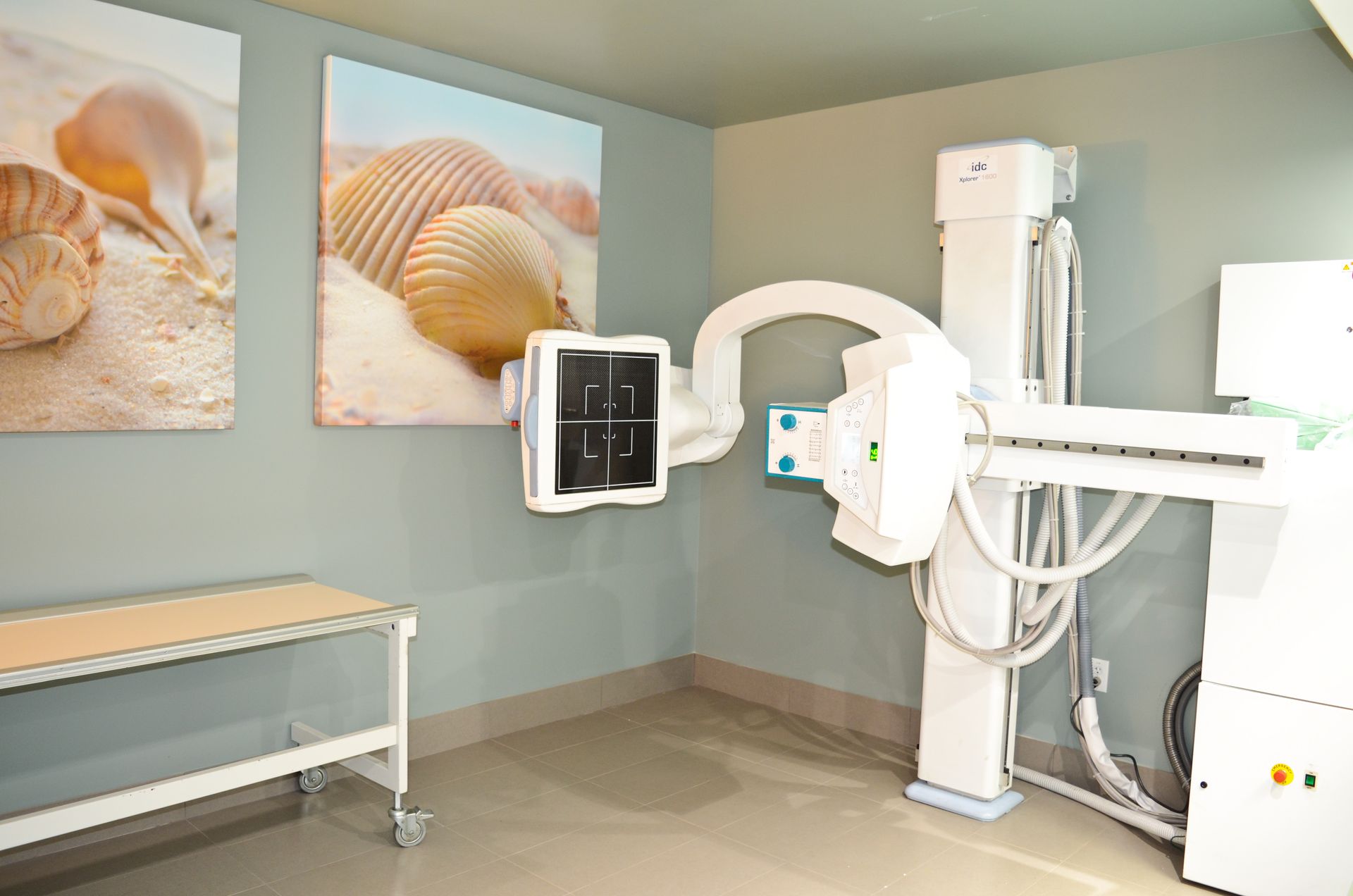 X-ray room with machine, exam table, beach-themed art, on gray walls.