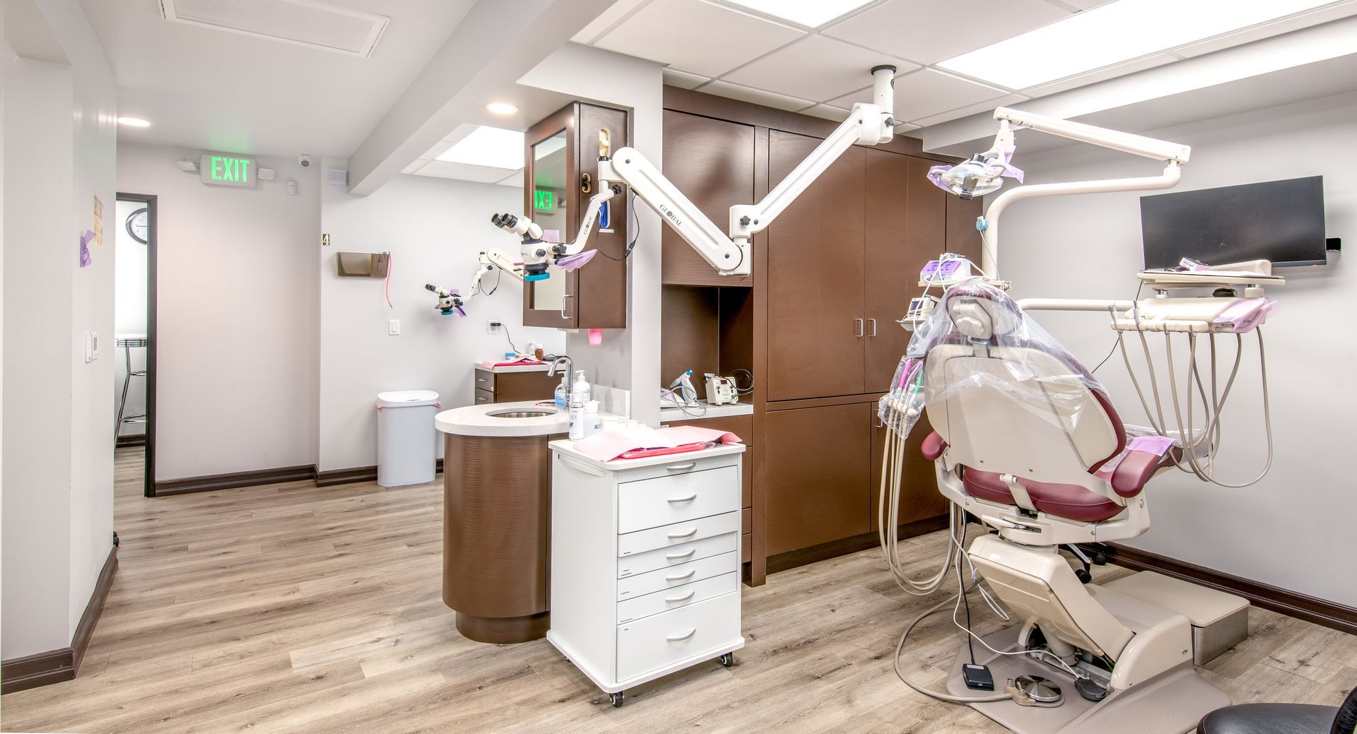 Dental office interior: dental chair, equipment, cabinets, light wood floor, exit door.