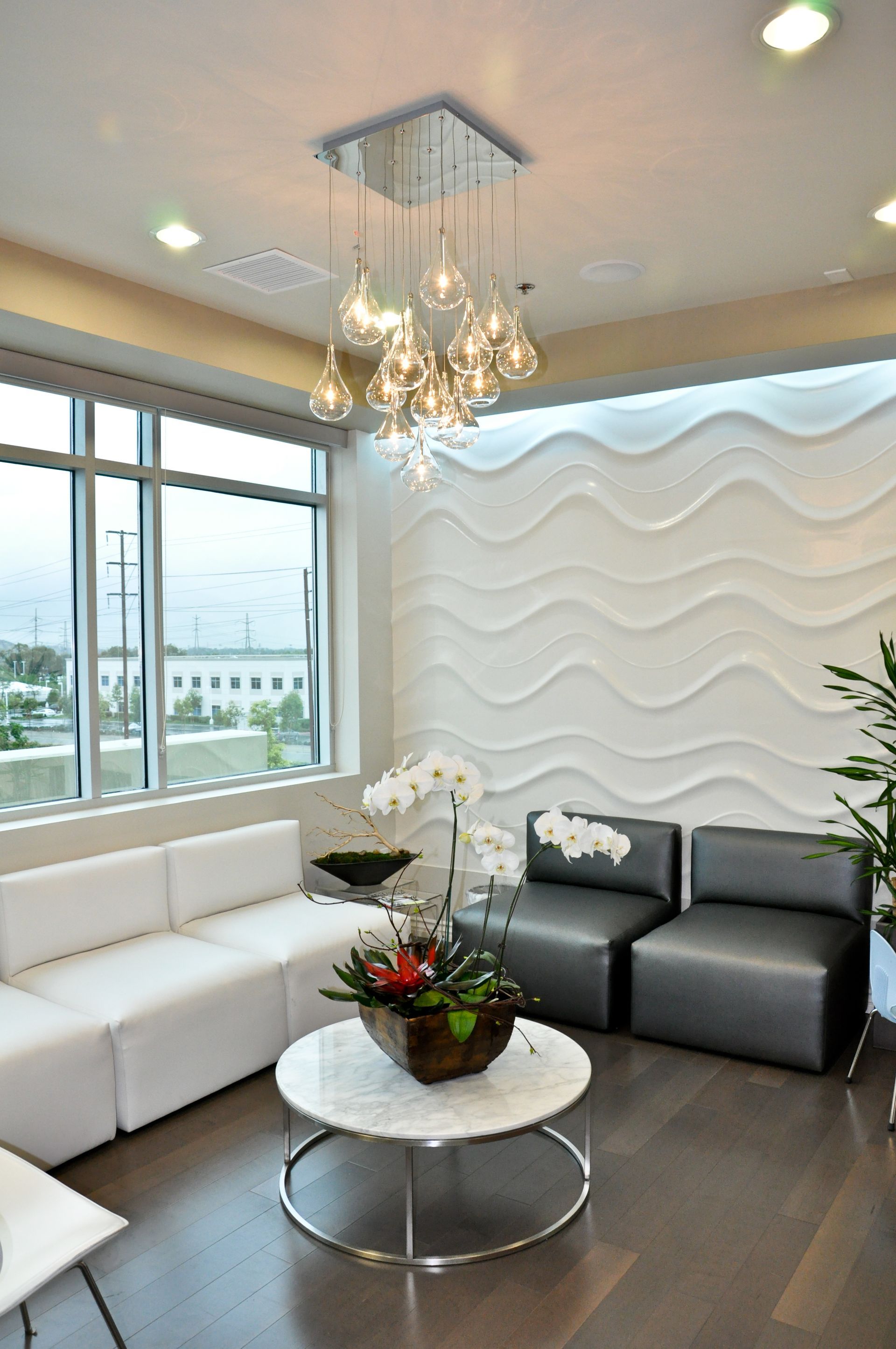 Modern waiting room with white sofa, gray chairs, and decorative wall. Glass globe chandelier hangs above a round table.