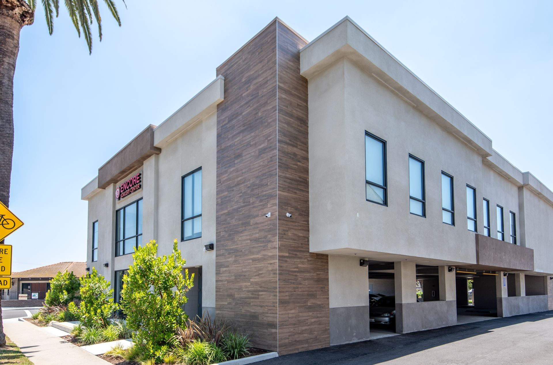 Two-story beige building with brown textured wall, blue windows, and palm tree against a clear sky.