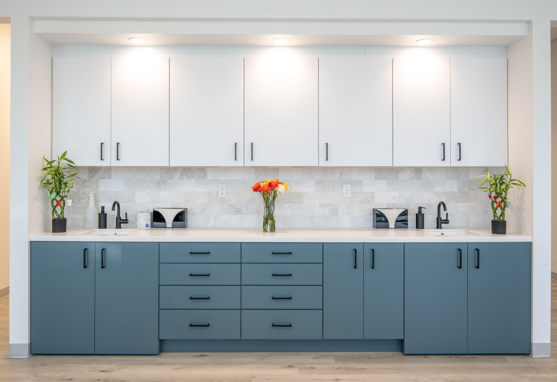 Modern office sink area with white and teal cabinets, marble tile backsplash, and flower vase.