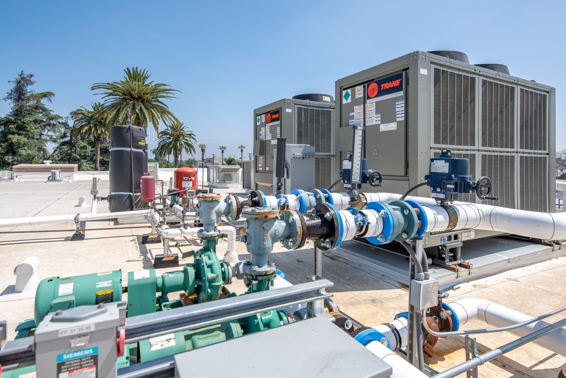 Rooftop with HVAC units, pipes, and pumps under a bright blue sky. Palm trees in the background.