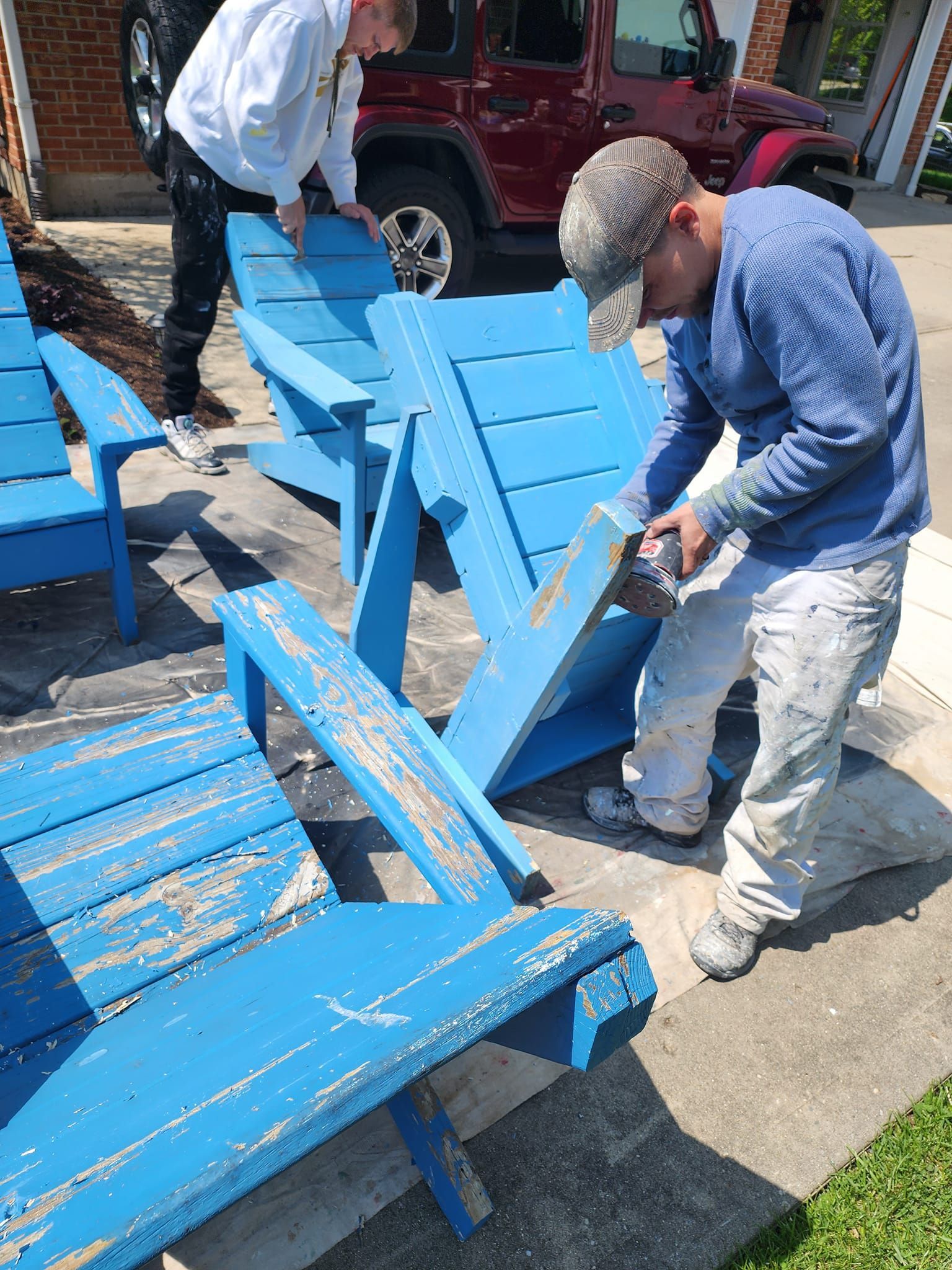 Two men are painting blue wooden chairs in a driveway.