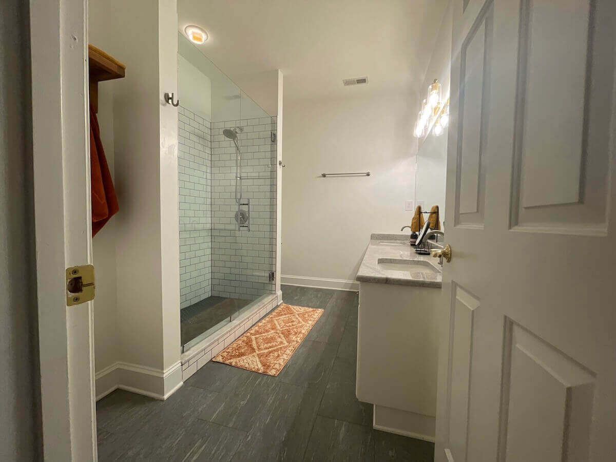 Bathroom with walk-in shower, white vanity, and gray flooring. A peach rug sits in front of the shower.