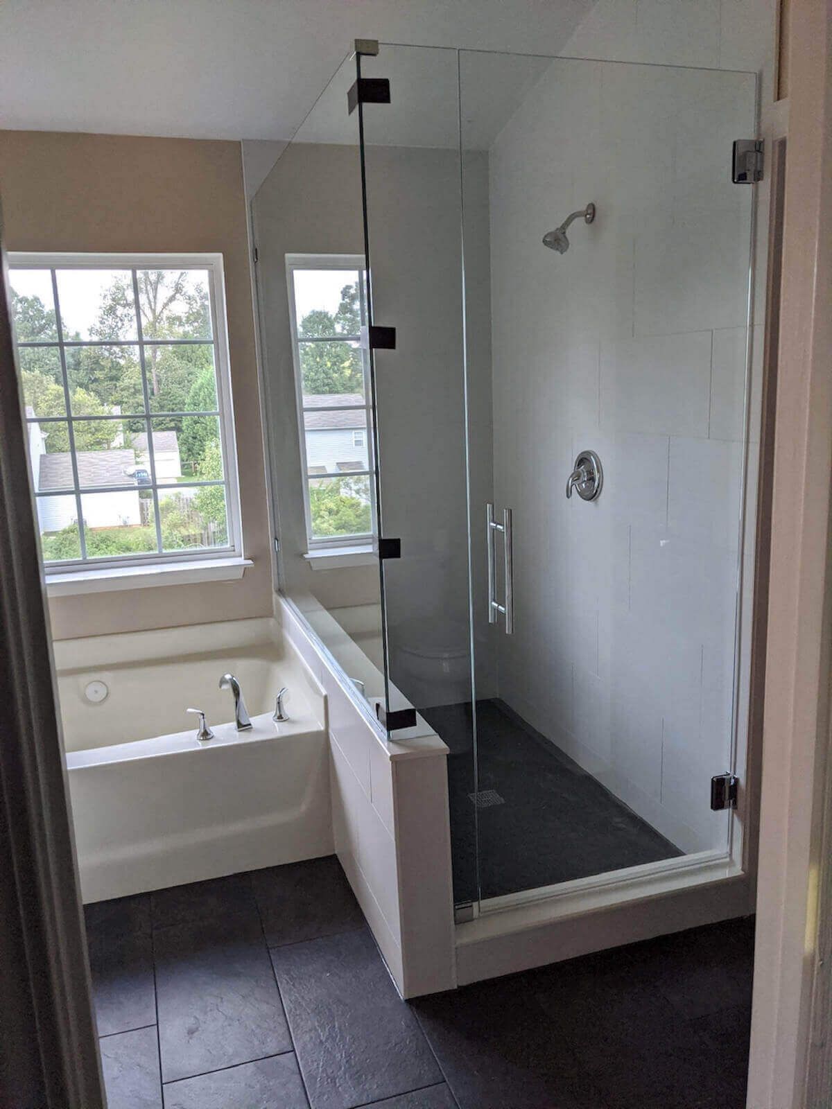 Modern bathroom with a glass shower enclosure next to a bathtub and window. Dark floor, white walls.