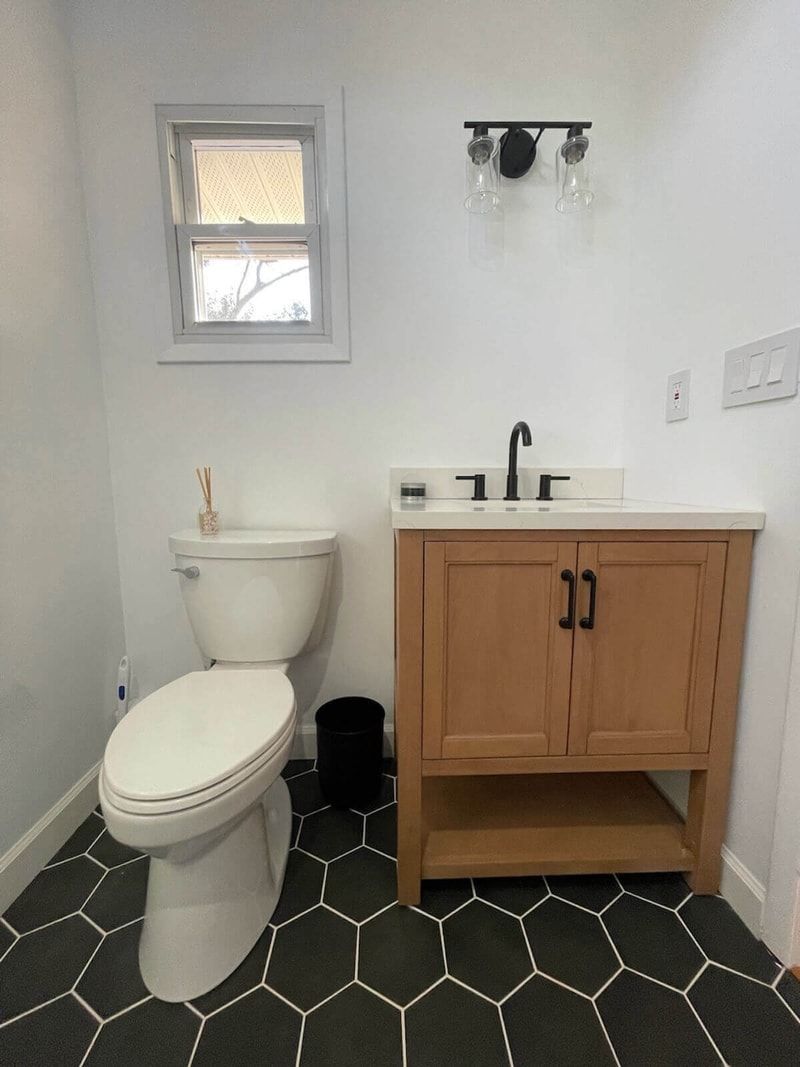 Bathroom with light wood vanity, white toilet, small window, black hexagonal floor tiles.