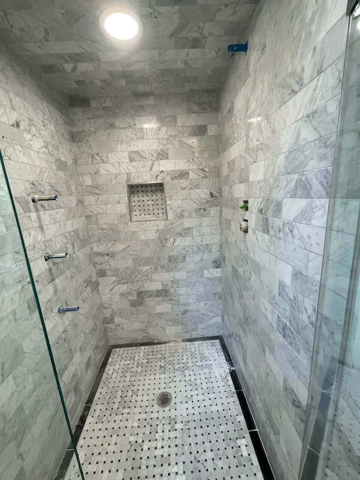 Marble-tiled shower with a niche and glass door. Stainless steel fixtures. Black border on the floor.