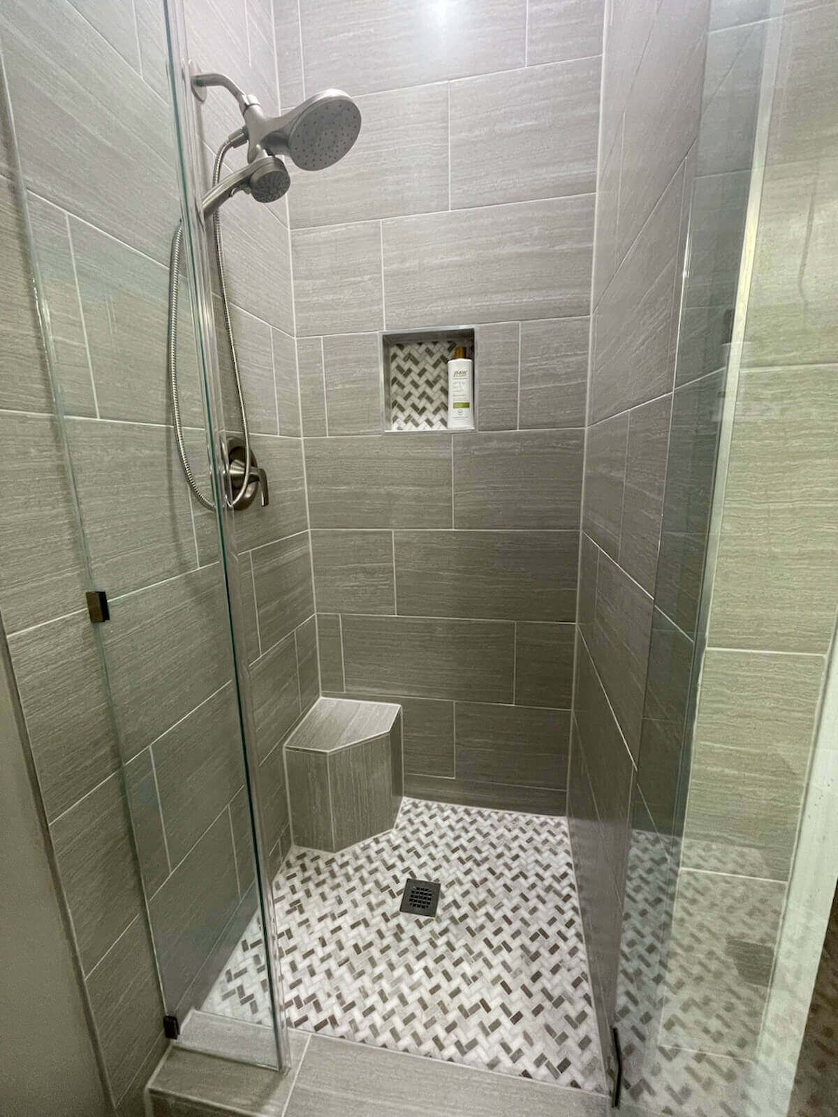 Gray tiled shower with glass door, bench, and patterned floor.