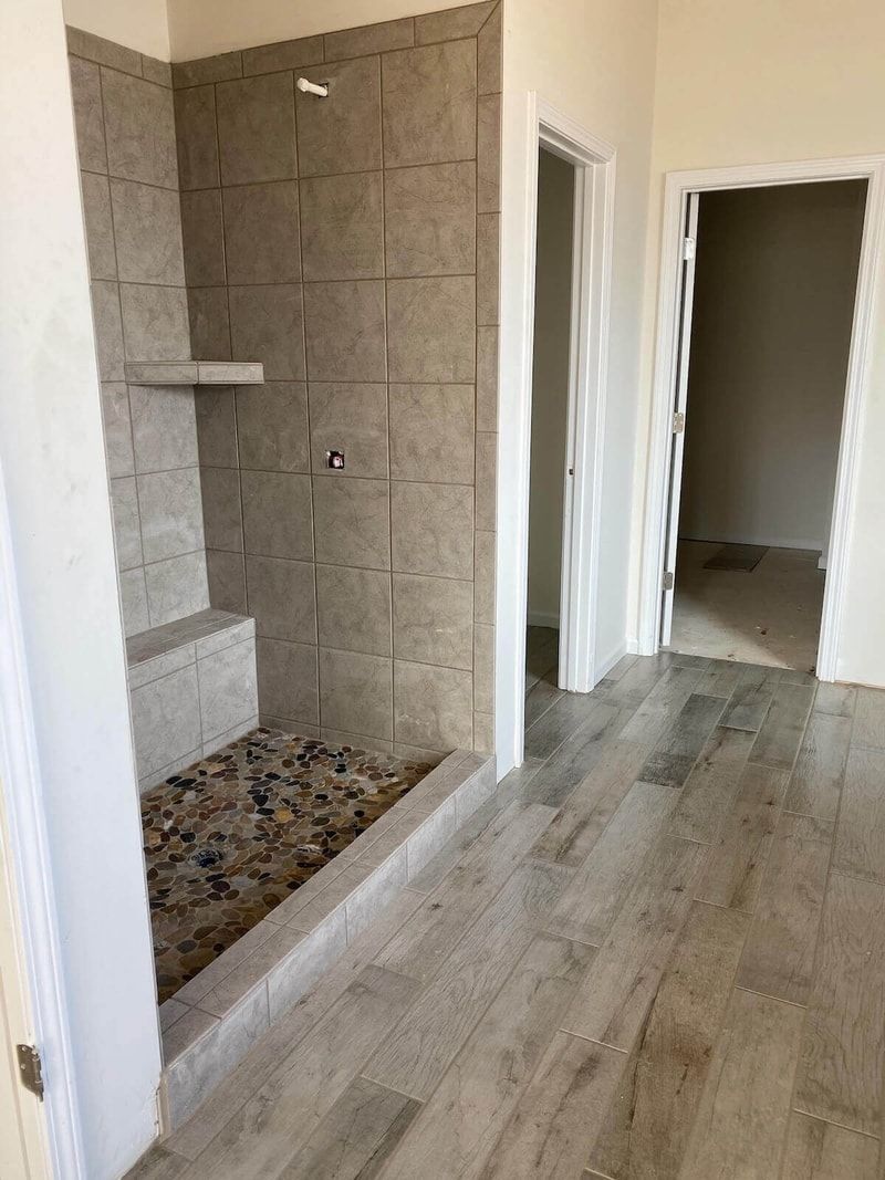 Bathroom with tiled shower, pebble floor, built-in seat, and two doorways. Gray tile and wooden floor.