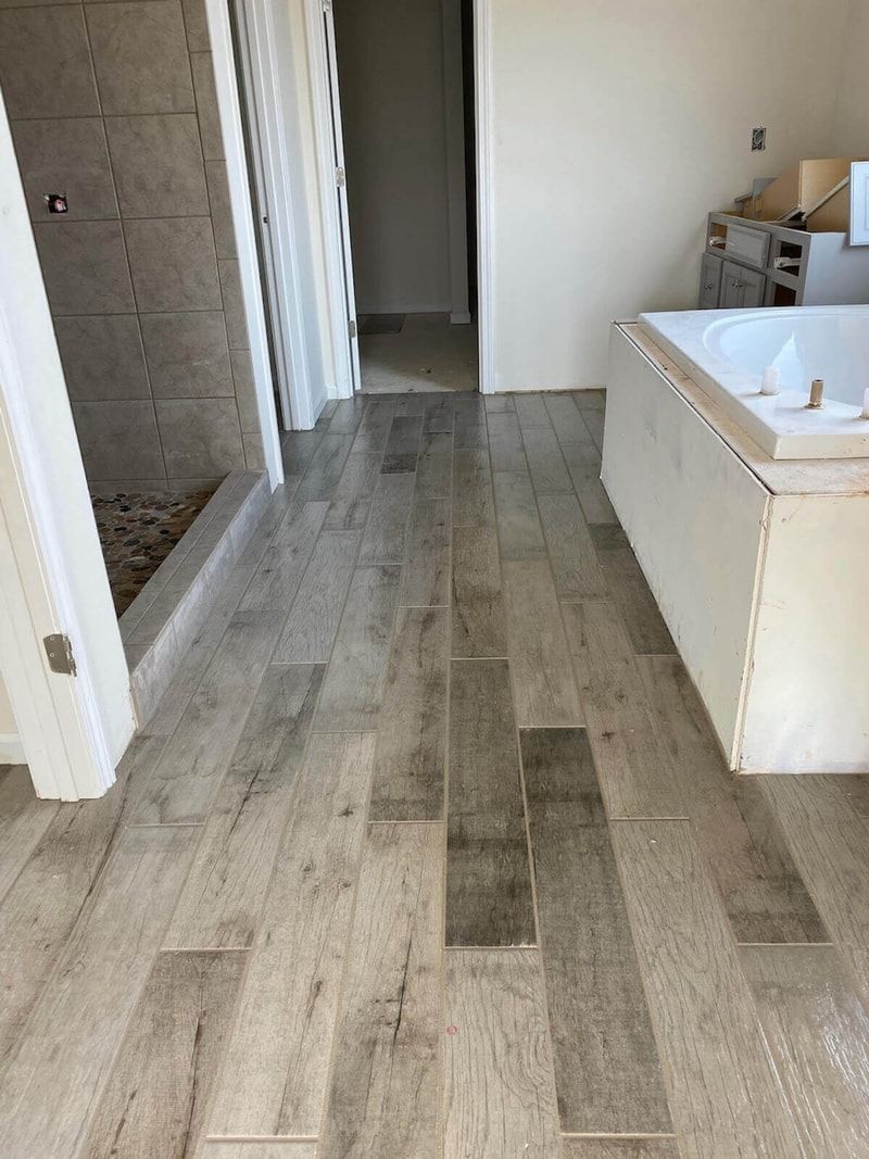 Bathroom with light wood-look floor tiles. Shower with gray tile visible. White bathtub and door frames.