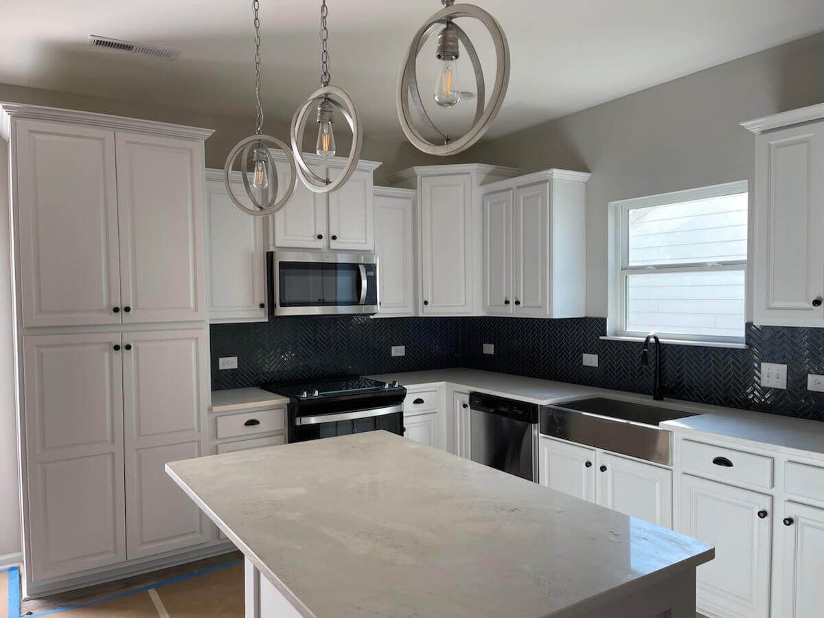 White kitchen with cabinets, island, black backsplash, stainless steel appliances, and pendant lights.