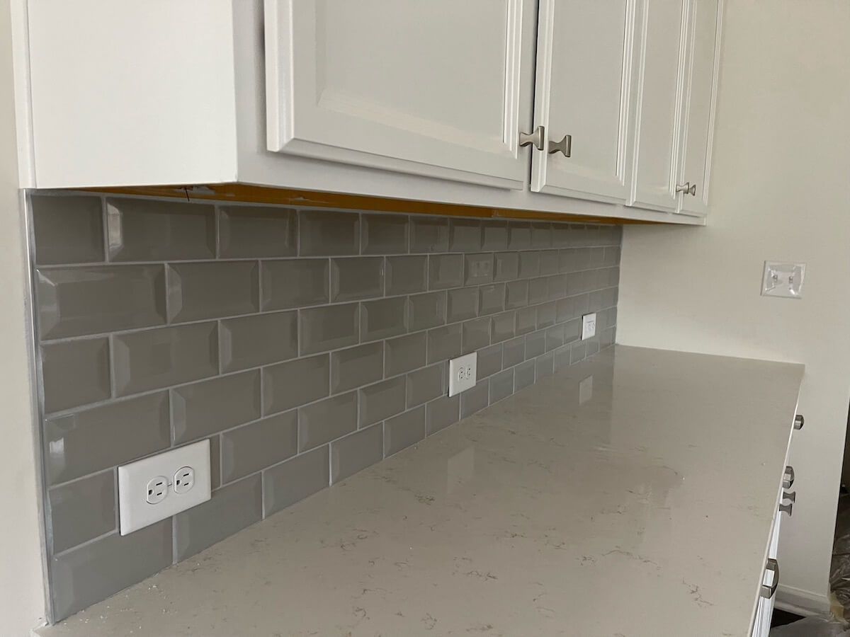 Gray tile backsplash and white cabinets in a kitchen, with countertops and electrical outlets.