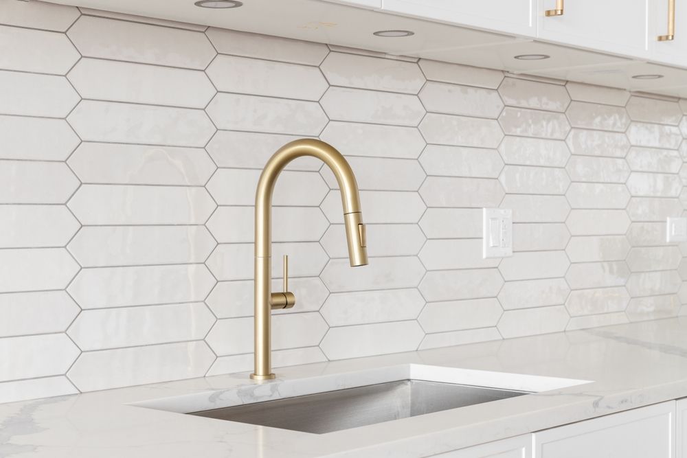 Gold faucet over a stainless steel sink, with white elongated hexagon tile backsplash.