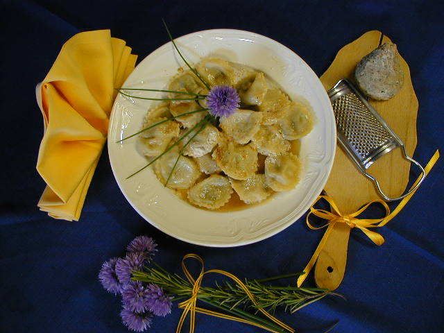 Ravioli impiattati con guarnizione di erbe aromatiche su un tavolo blu, affiancati da tovaglioli gialli e una grattugia con utensili.