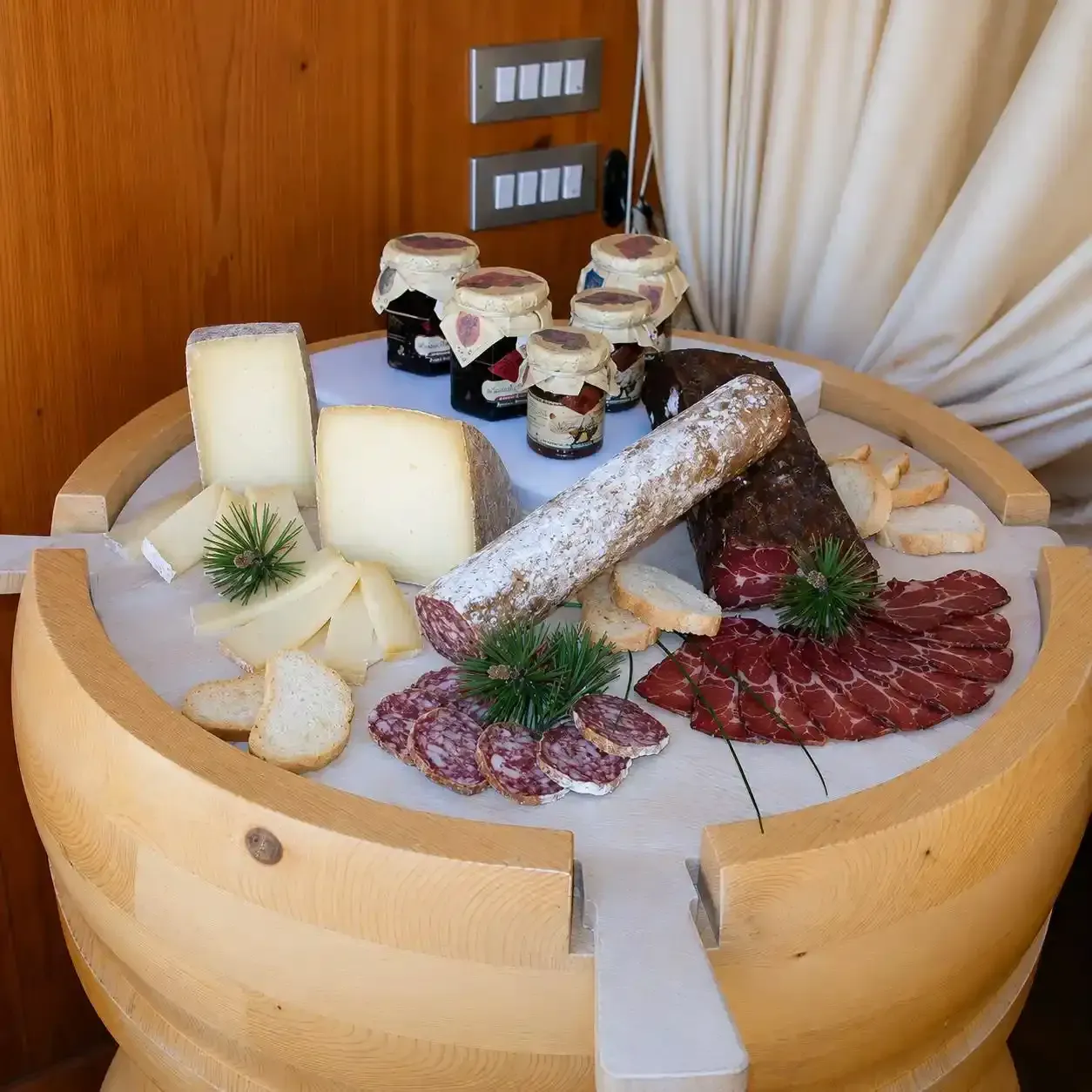 Piatto di salumi e formaggi su un tavolo ricavato da una botte di legno con salumi, spicchi di formaggio e vasetti di crema spalmabile.
