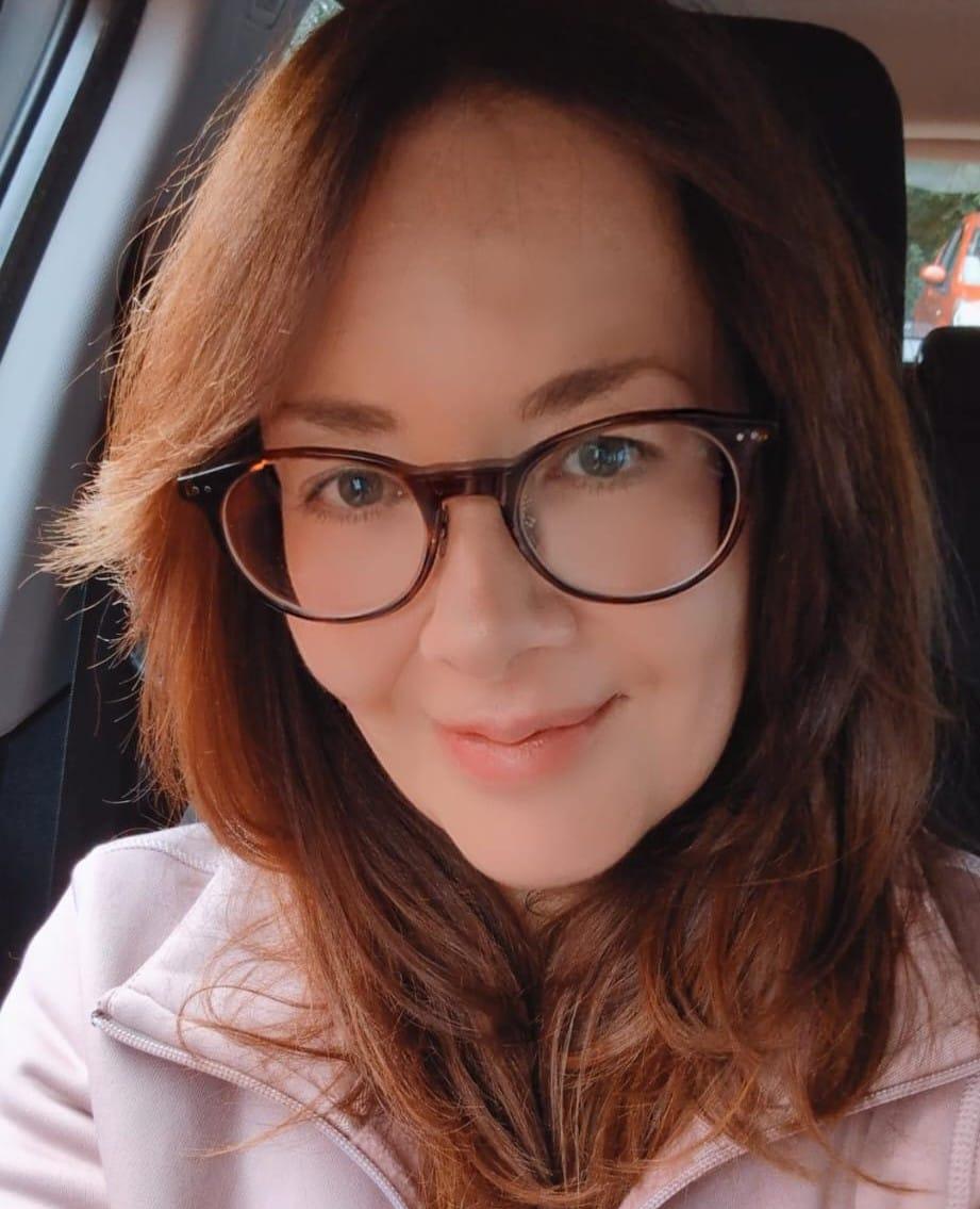 Eine Person mit dunkelumrandeter Brille und hellrosa Jacke posiert für ein Selfie in einem Fahrzeug.