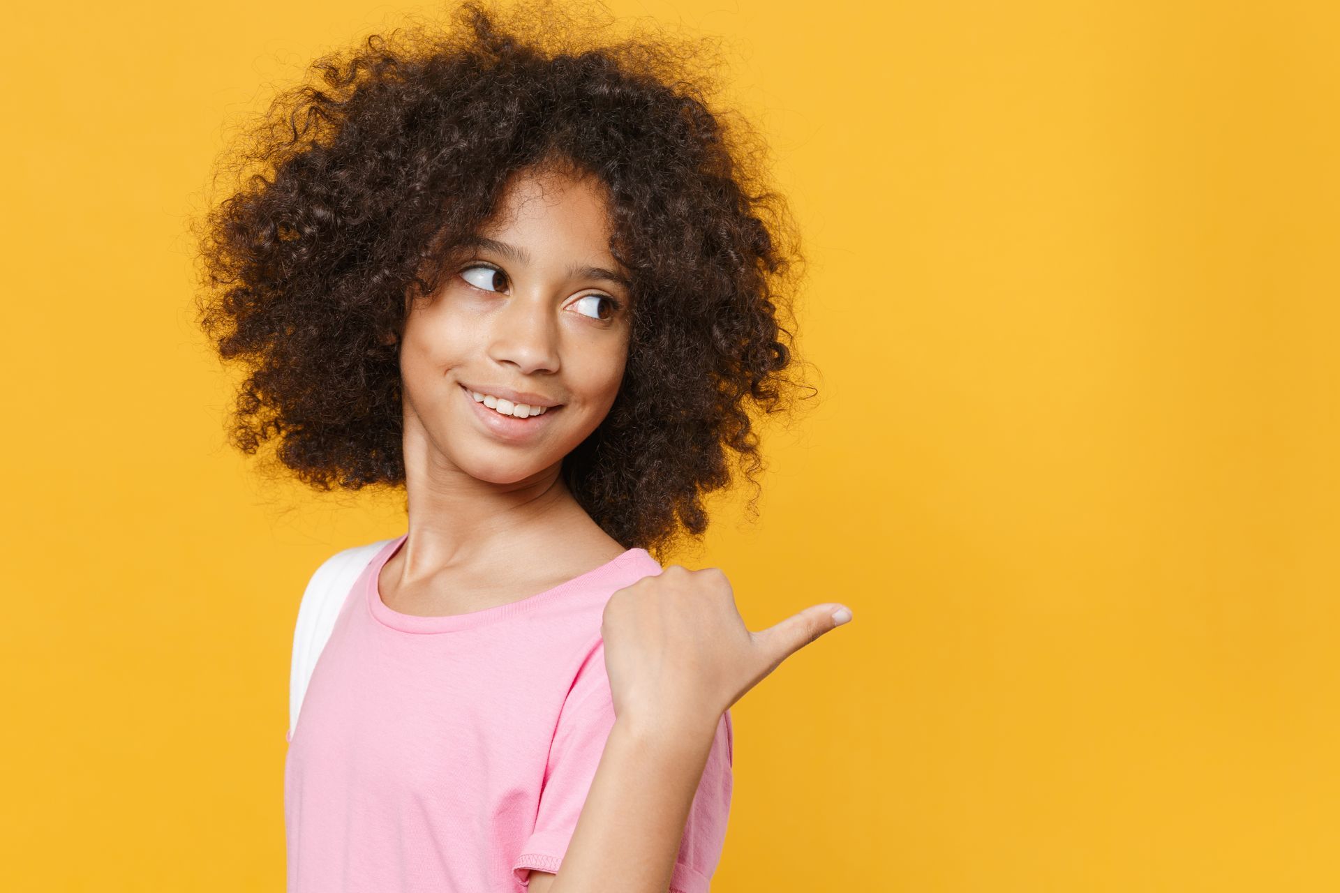 Girl with curly hair in pink shirt points right, smiles against yellow background.