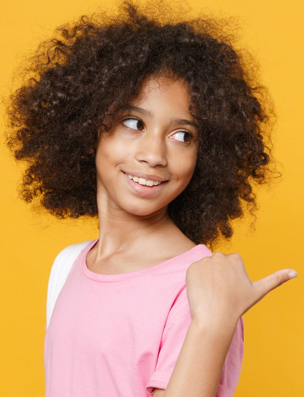 Girl with curly hair, pink shirt, smiling, pointing to the right against a yellow background.