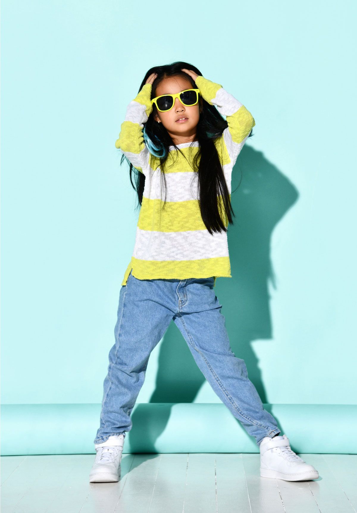 A little girl wearing sunglasses and a striped sweater is standing on a blue background.